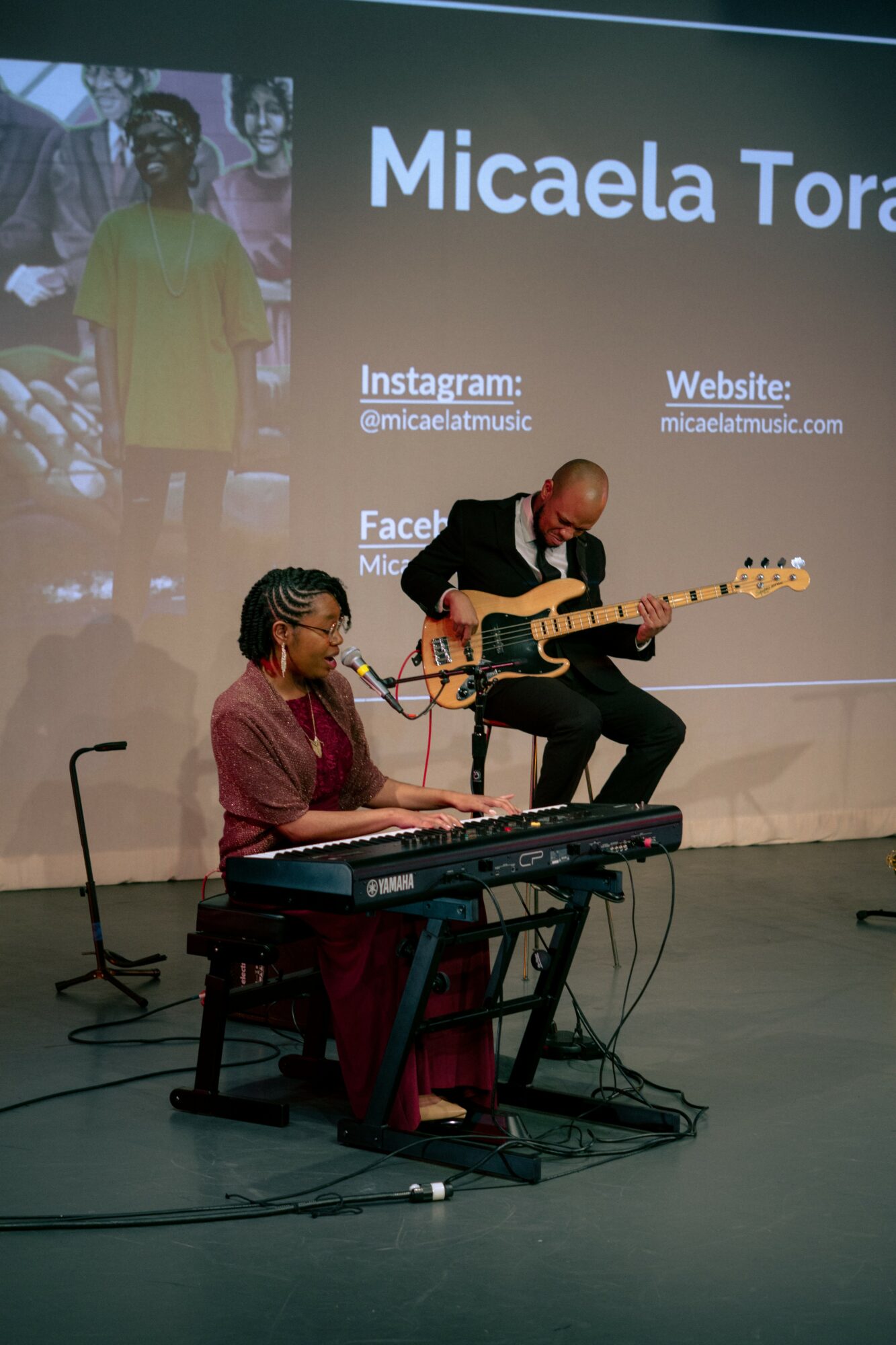 Two musicians perform on stage, one playing keyboard and the other playing guitar, with a screen displaying text behind them.