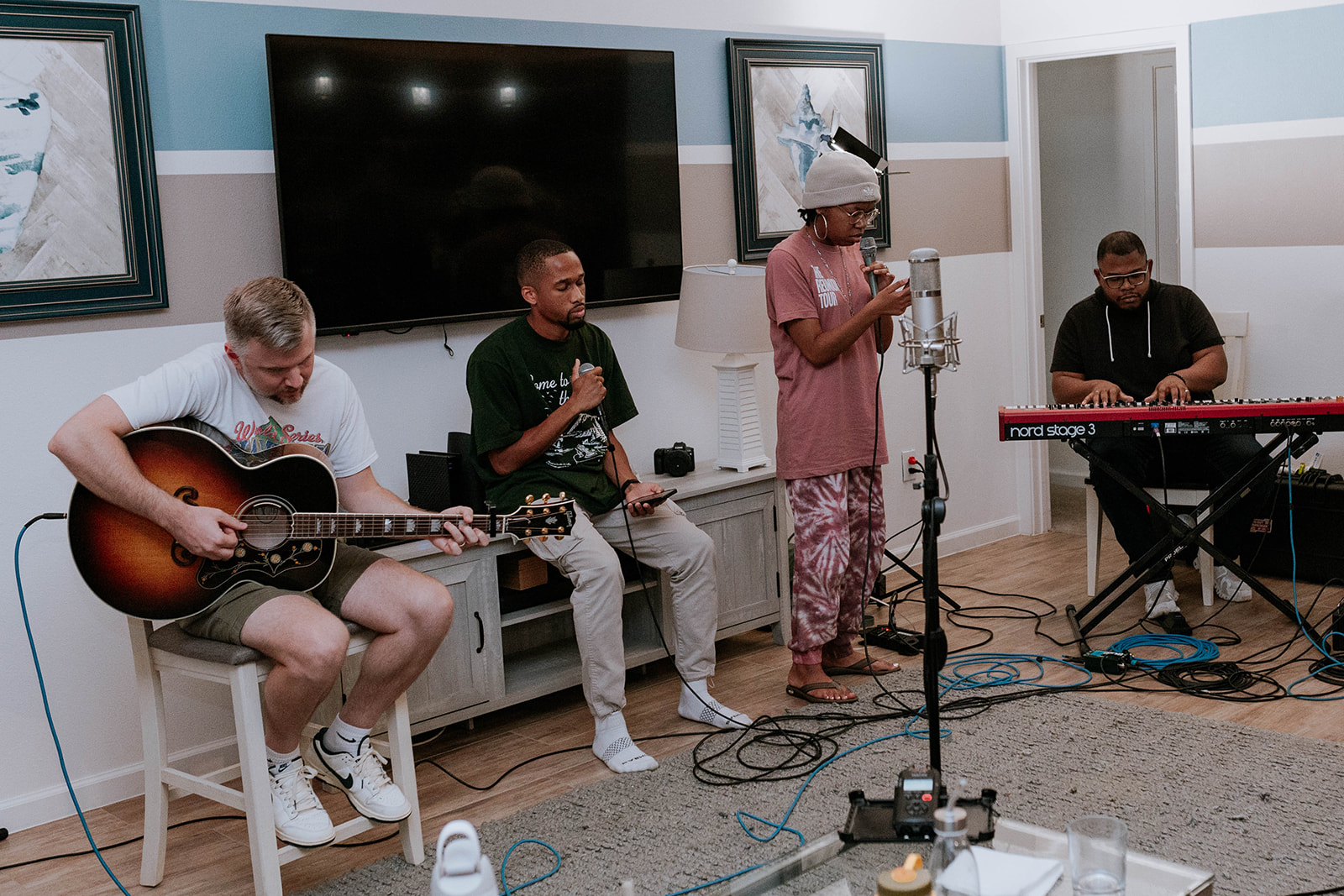 Four musicians perform in a room with a large screen, paintings, and a keyboard, playing instruments and singing.