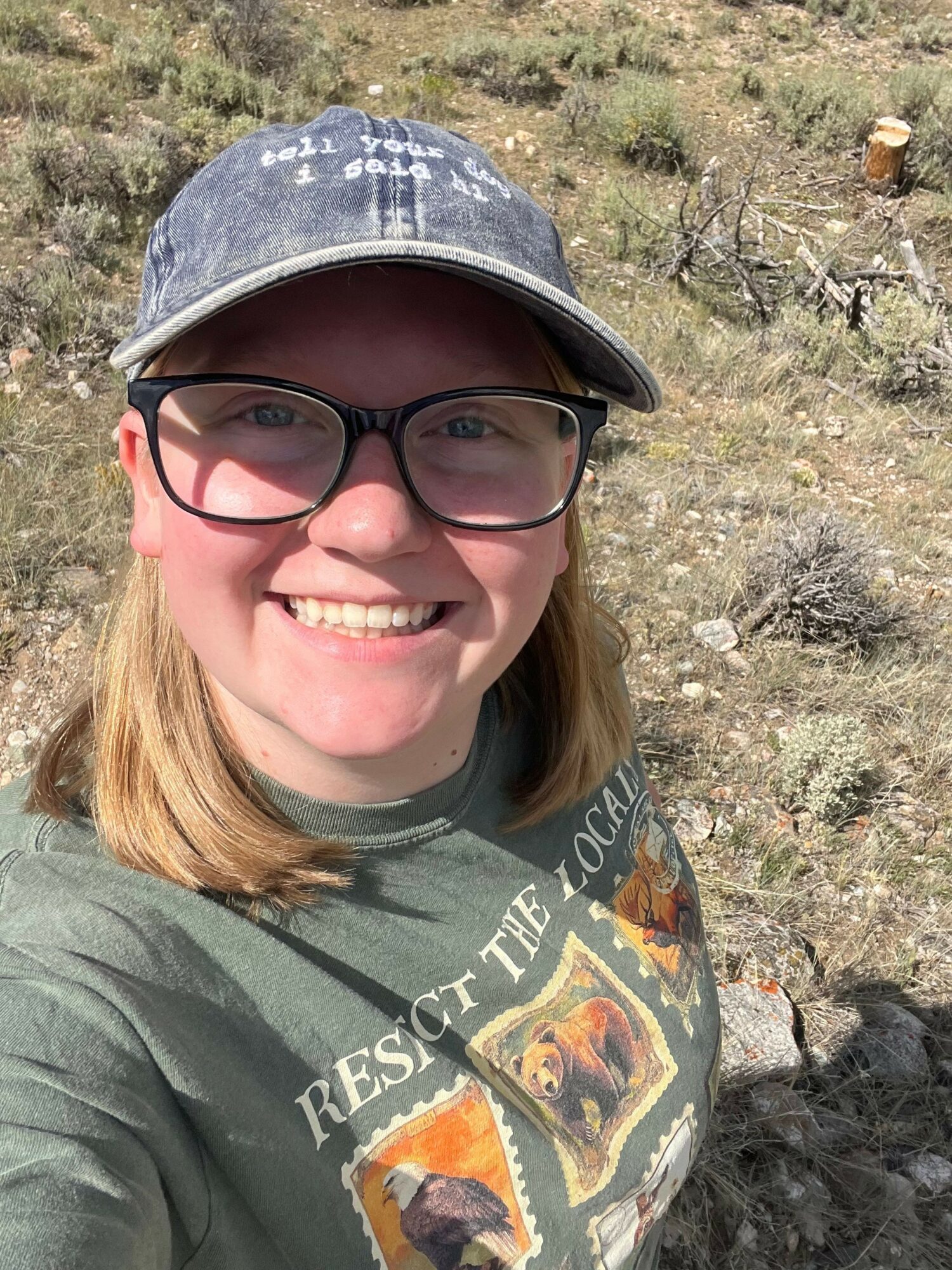 Smiling person wearing glasses and a cap outdoors, with rocky terrain and vegetation in background.