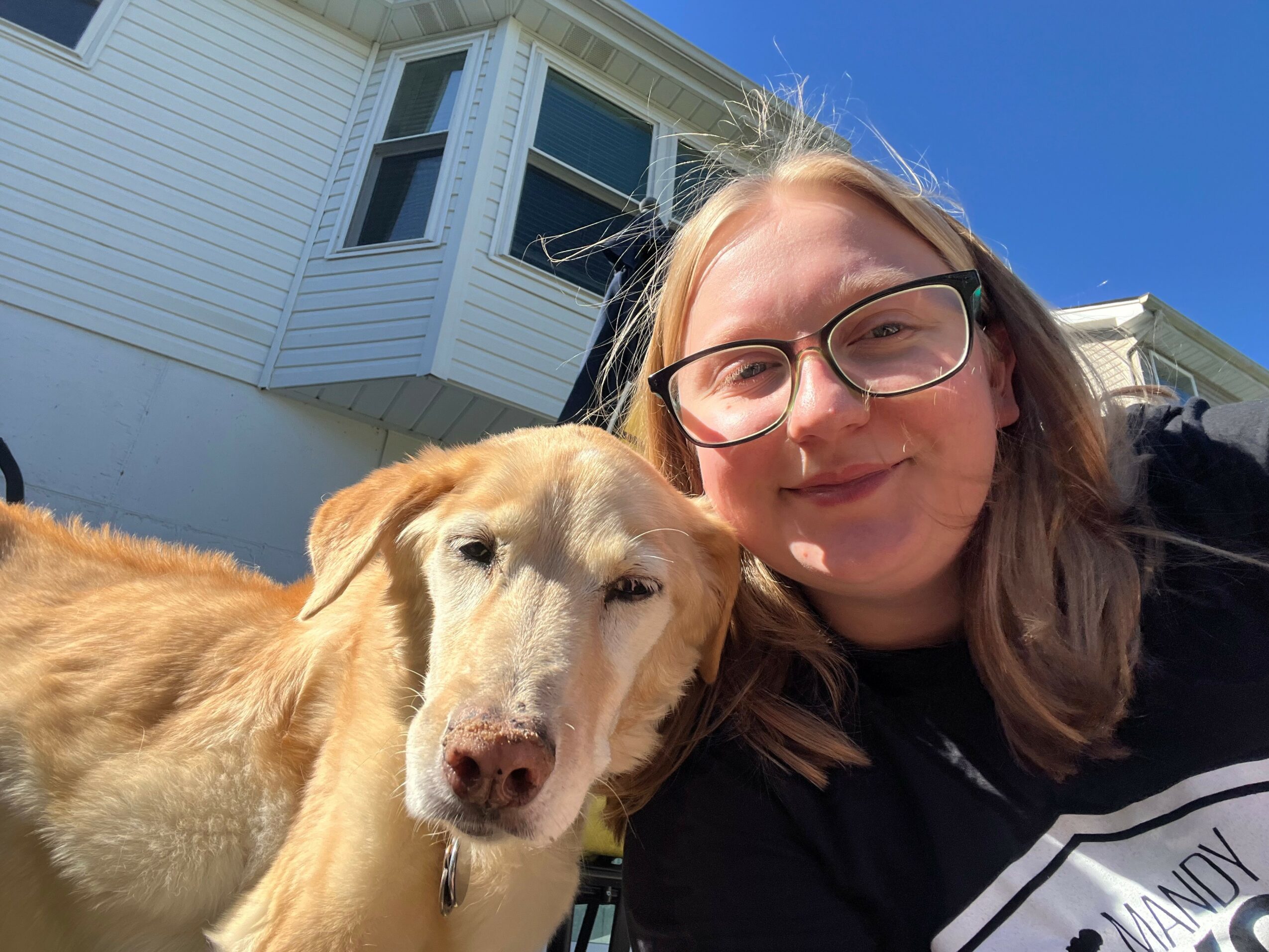 Person with glasses smiling next to a light-colored dog outdoors in front of a white house with blue sky.