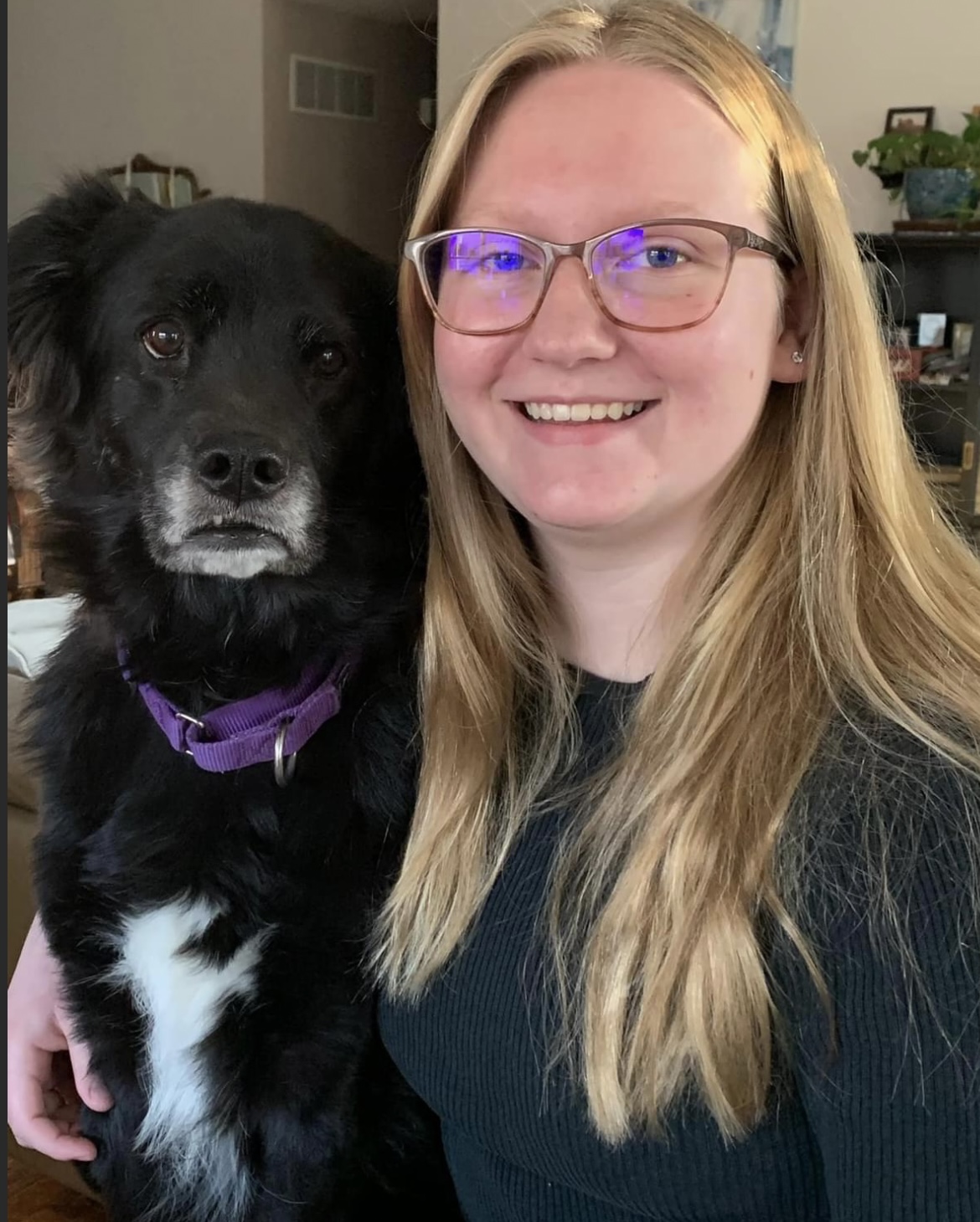 A smiling woman with long blonde hair and glasses poses with a black dog wearing a purple collar indoors.