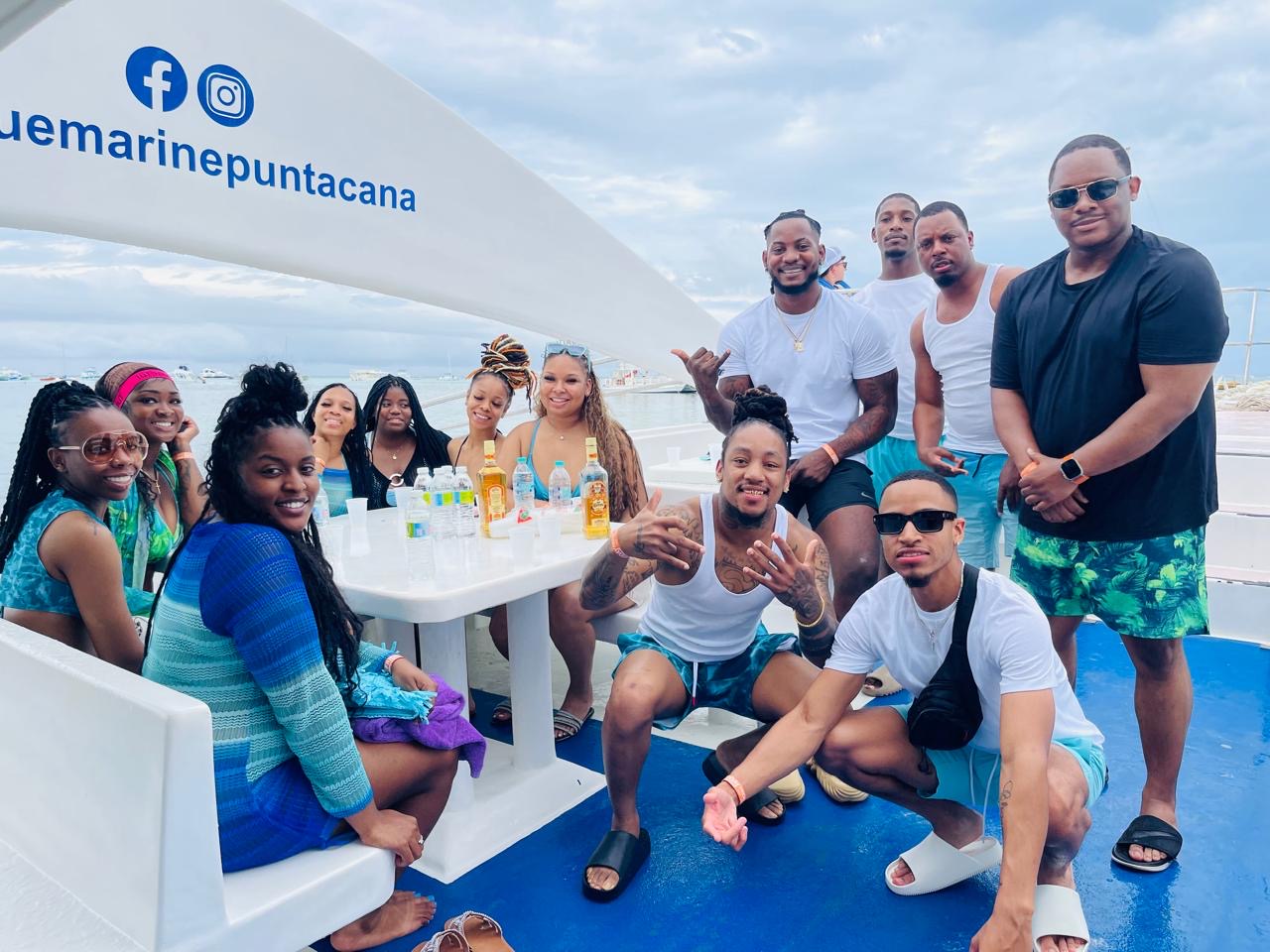 Group of people enjoying a boat trip with a table of drinks, some sitting and some standing, on a boat with a blue sky background.