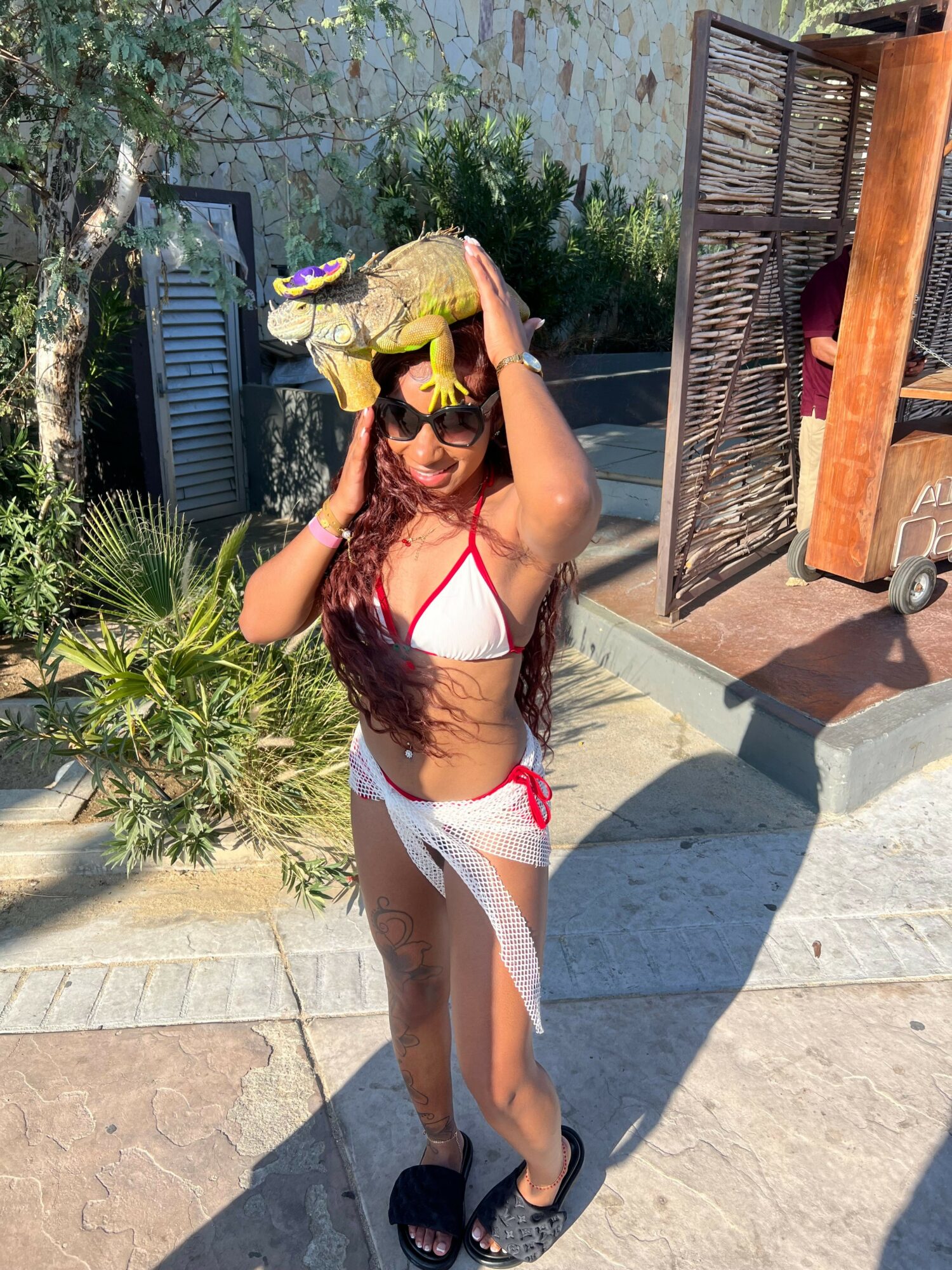 Woman in swimsuit and flip-flops standing outdoors, holding her hair, with plants and a wooden structure in background.