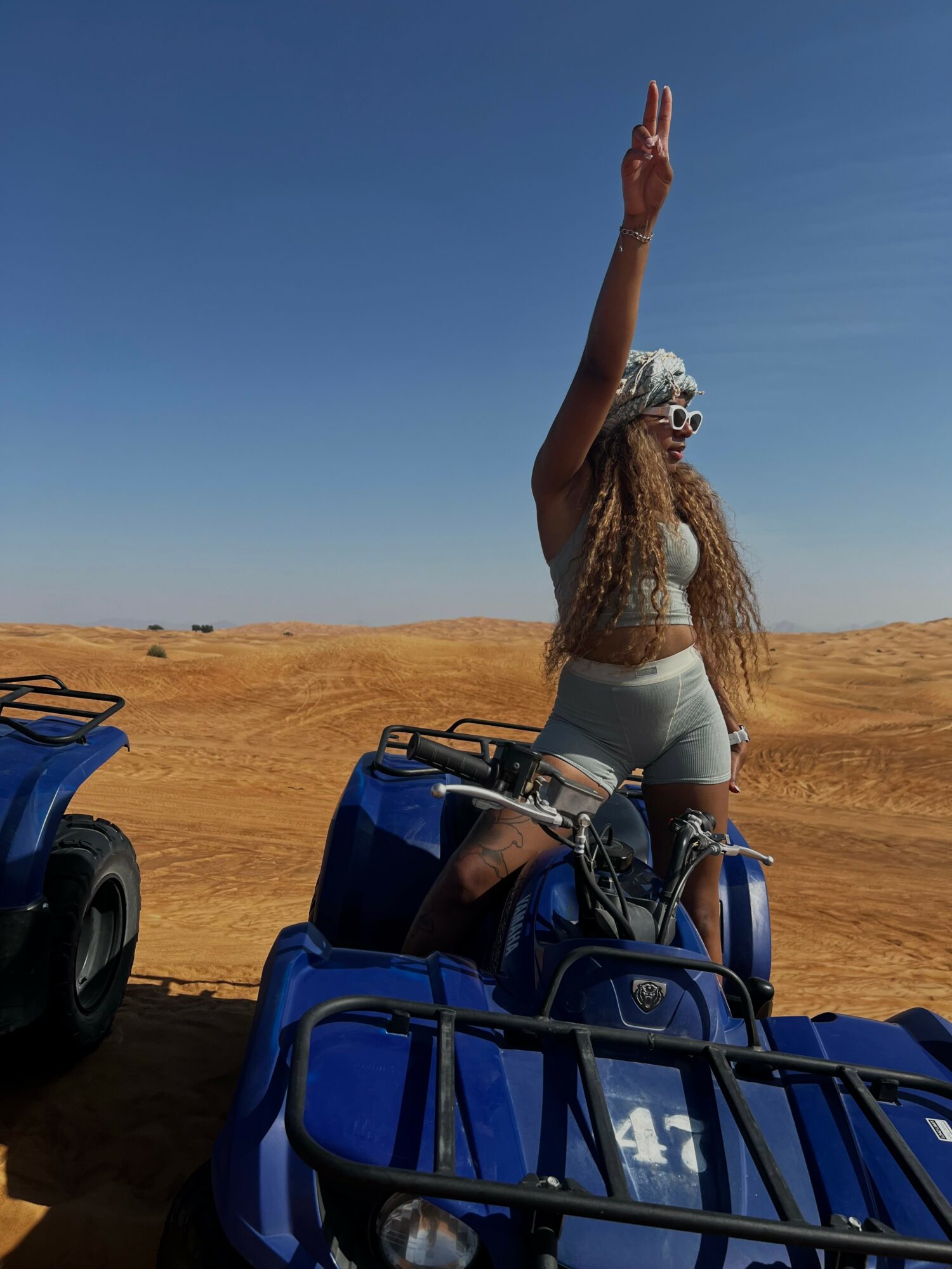 Person standing on an all-terrain vehicle in a desert, raising one arm, with clear blue sky above.