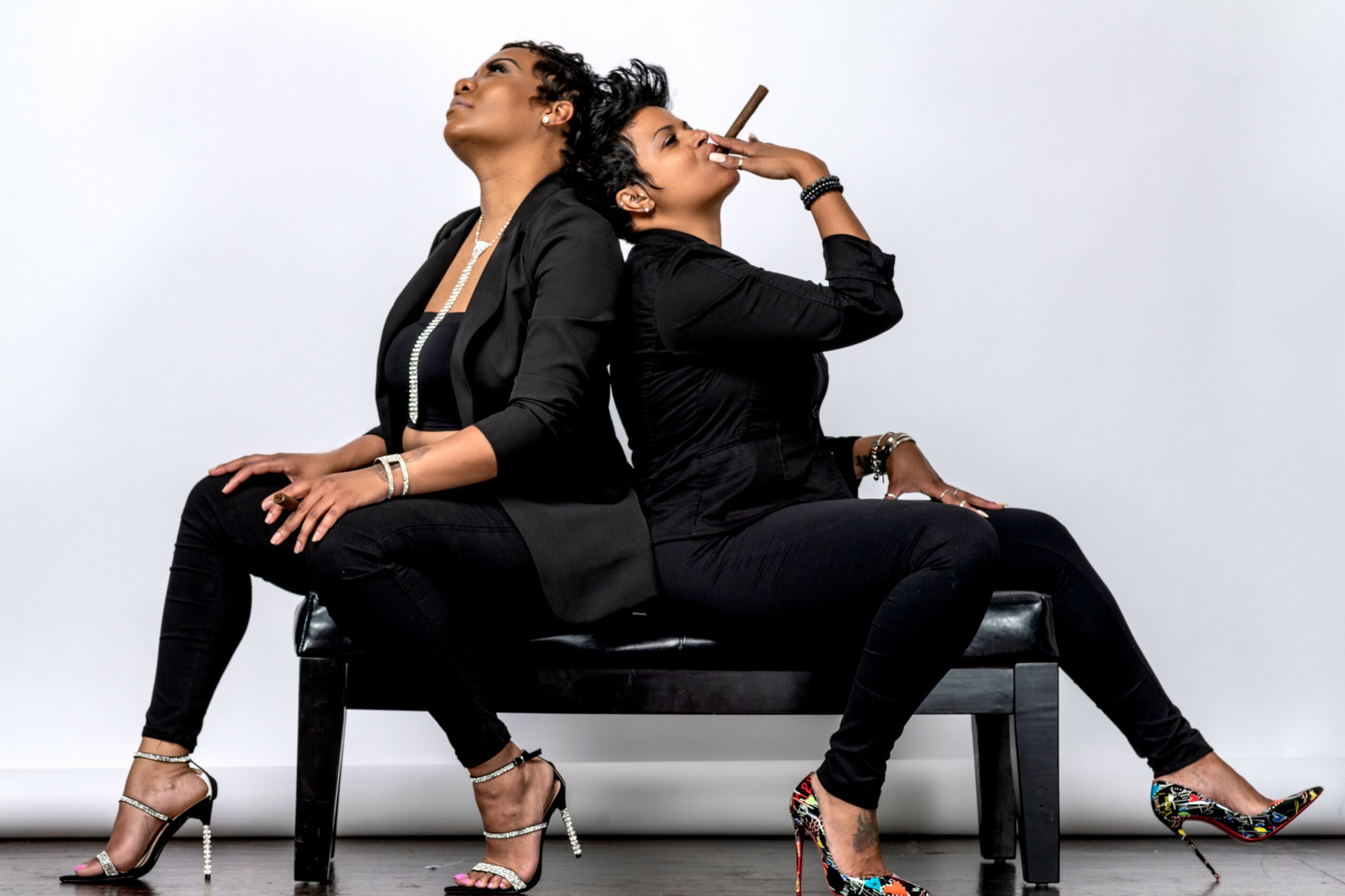 Two women sit back-to-back on a black bench, one smoking a cigarette, both dressed in black, with high heels, against a plain white background.
