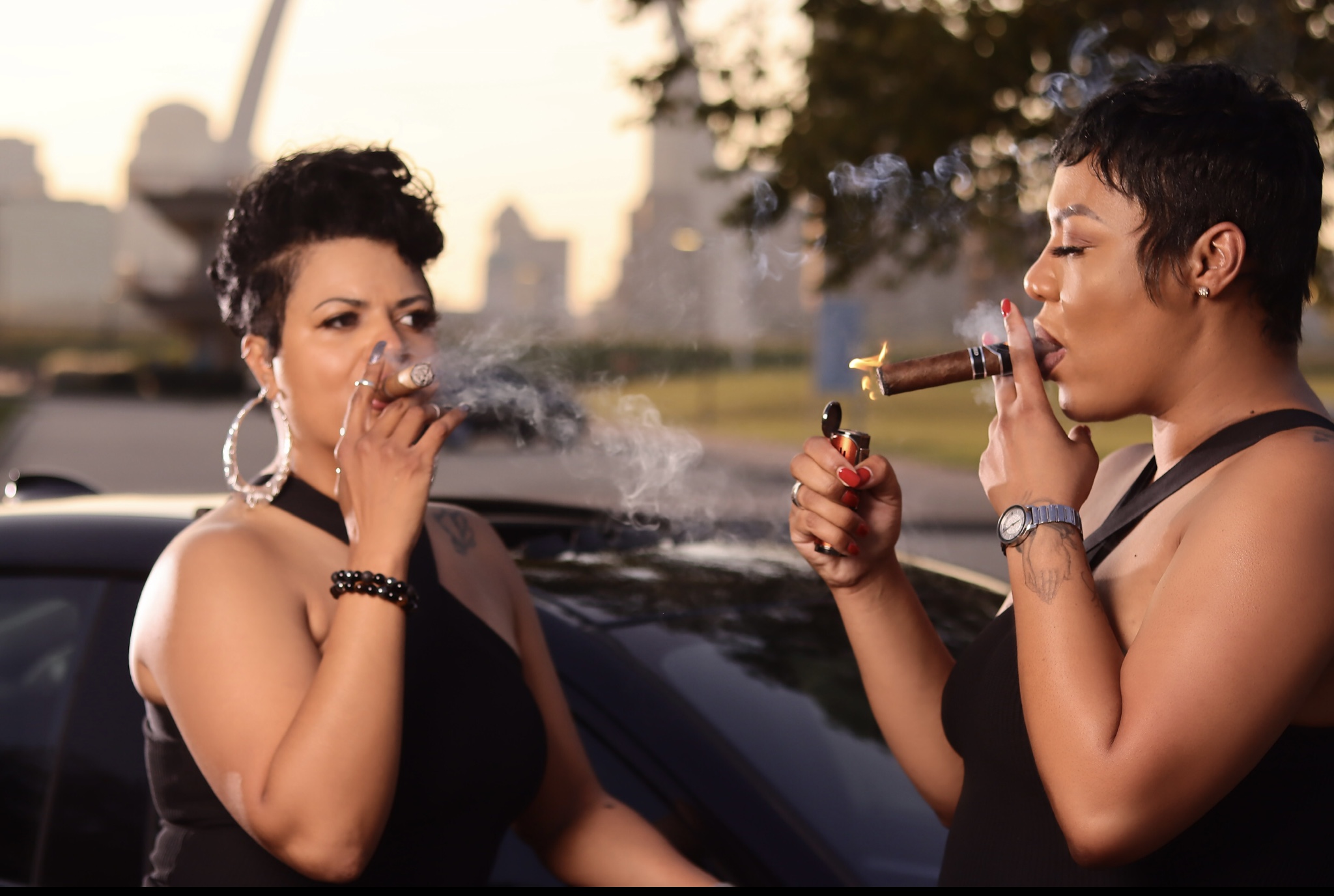 Two women with short hair and earrings, one drinking and the other smoking a cigar, outdoors near a car.