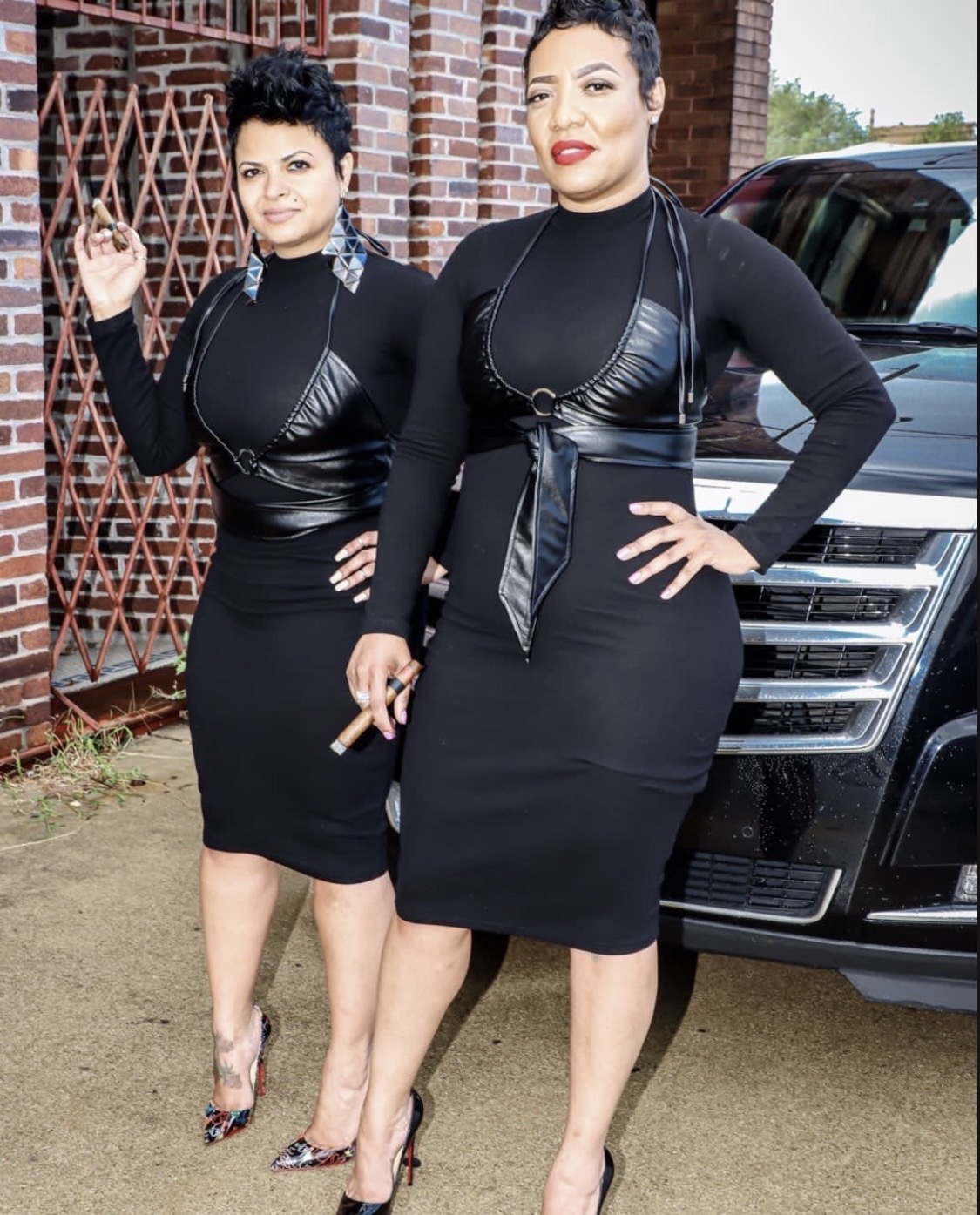 Two women in black dresses and high heels standing outdoors near a black vehicle and brick wall.