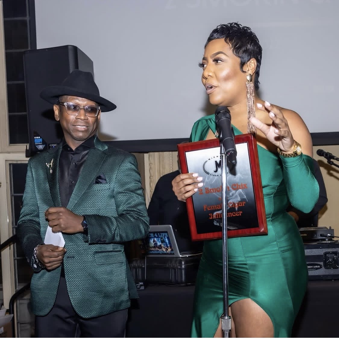 A woman in a green dress speaking into a microphone, holding a framed award, with a man in a suit and hat nearby.