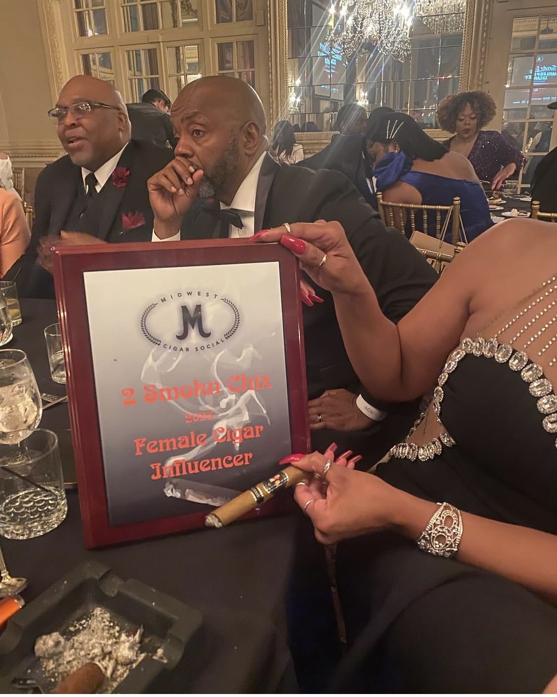 People seated at a table during an event, with a woman holding a framed award or certificate, and a woman in a black dress with jewelry.