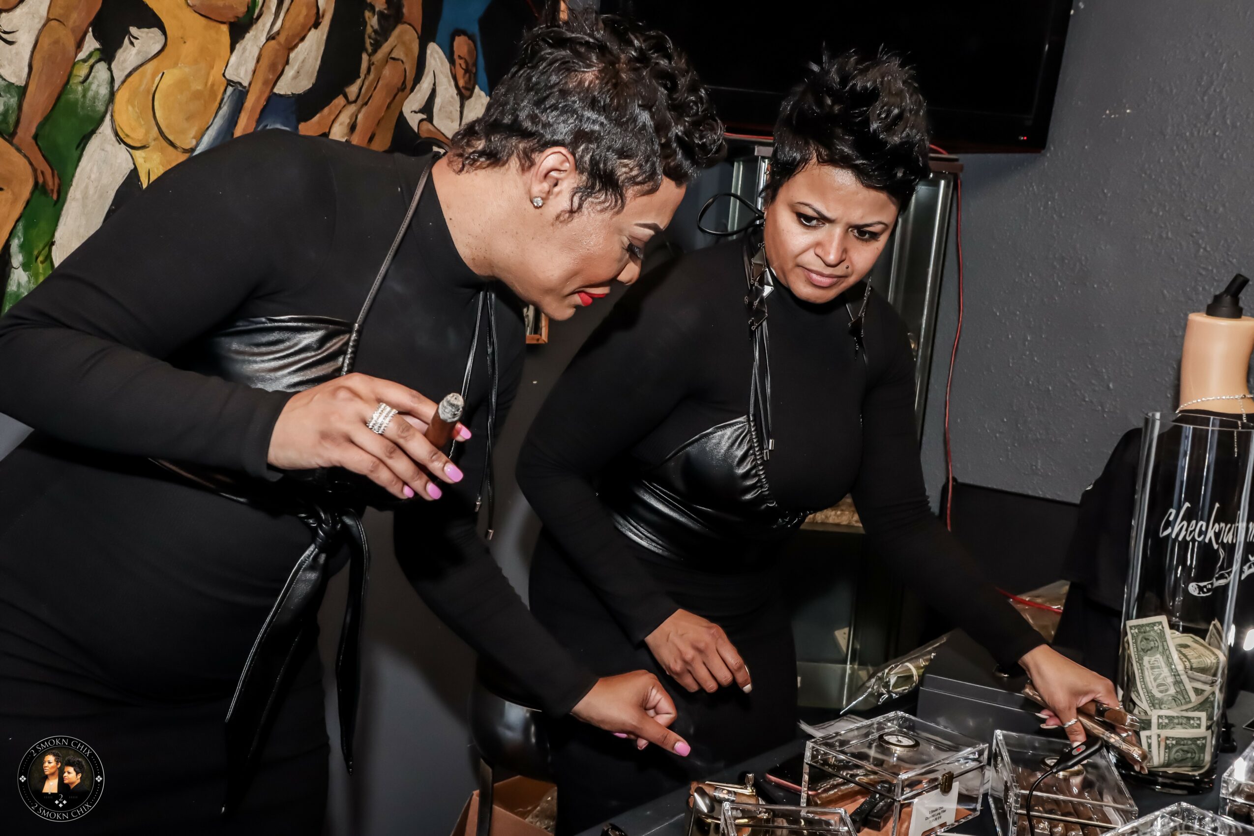 Two women in black outfits are looking at a display case with jewelry or accessories, in a dimly lit setting.