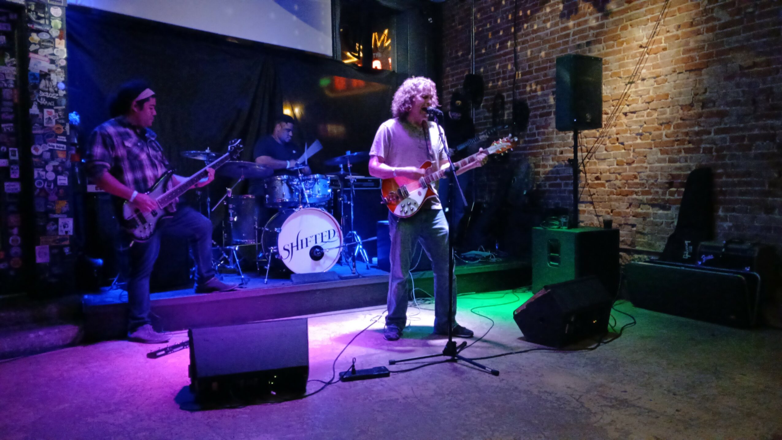 Band performing live on stage with four musicians, colorful lighting, brick wall background, and musical equipment.