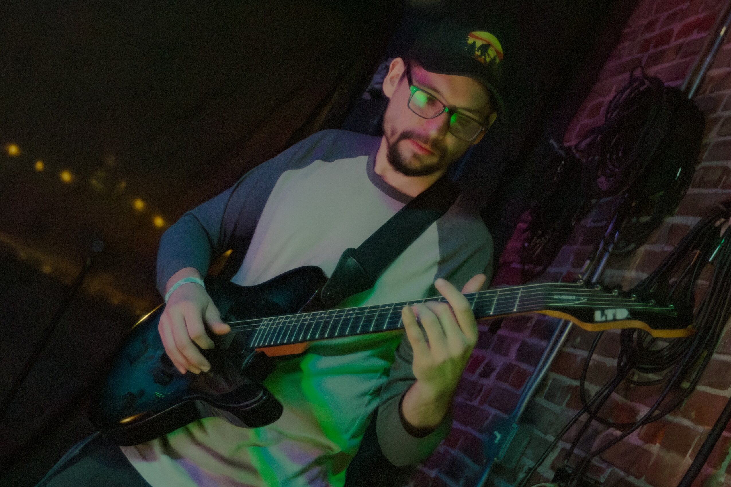 Man with glasses and beard playing an electric guitar in a dimly lit room with brick wall background.