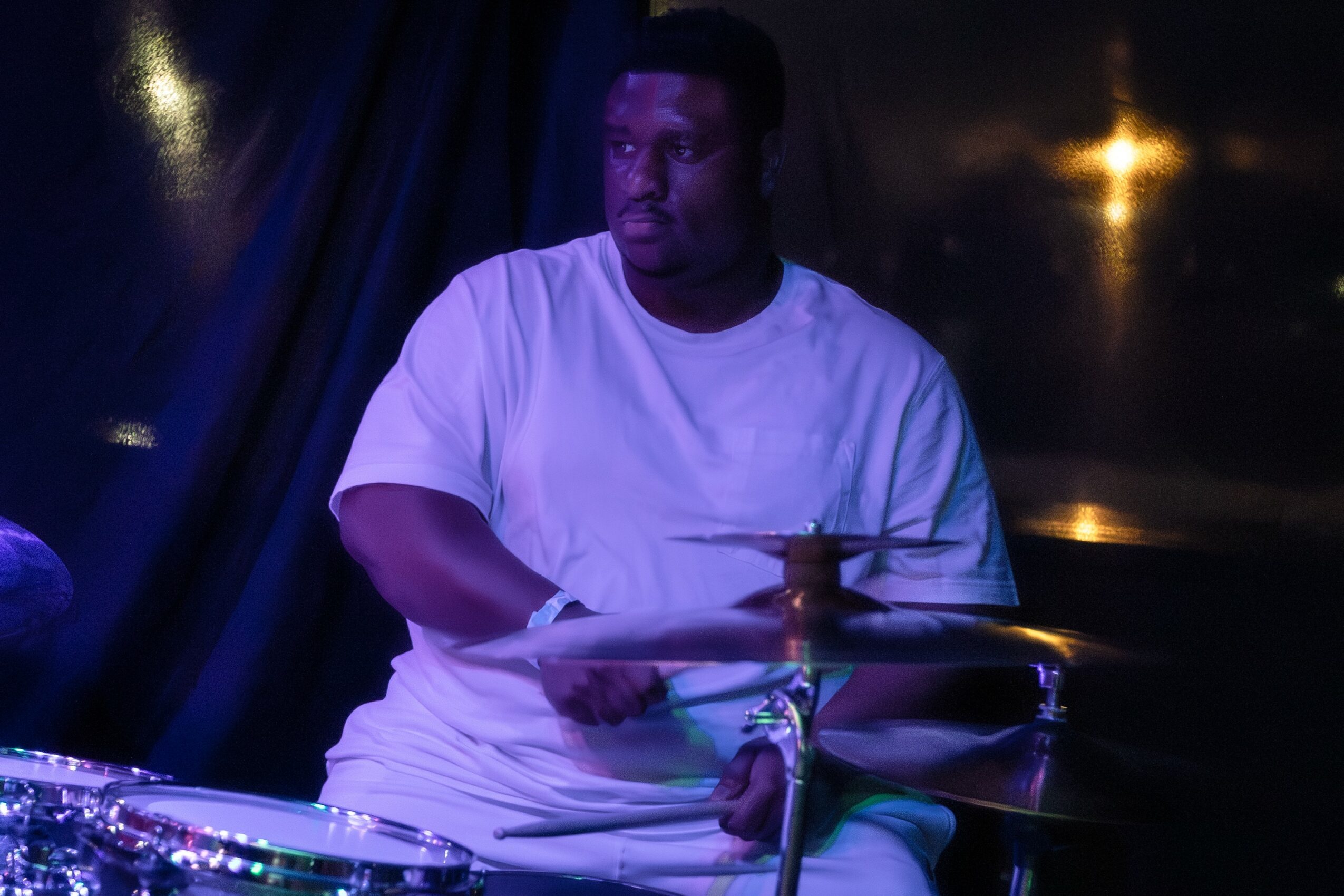 Person playing drums on stage, wearing a white shirt, with dark background and stage lighting.