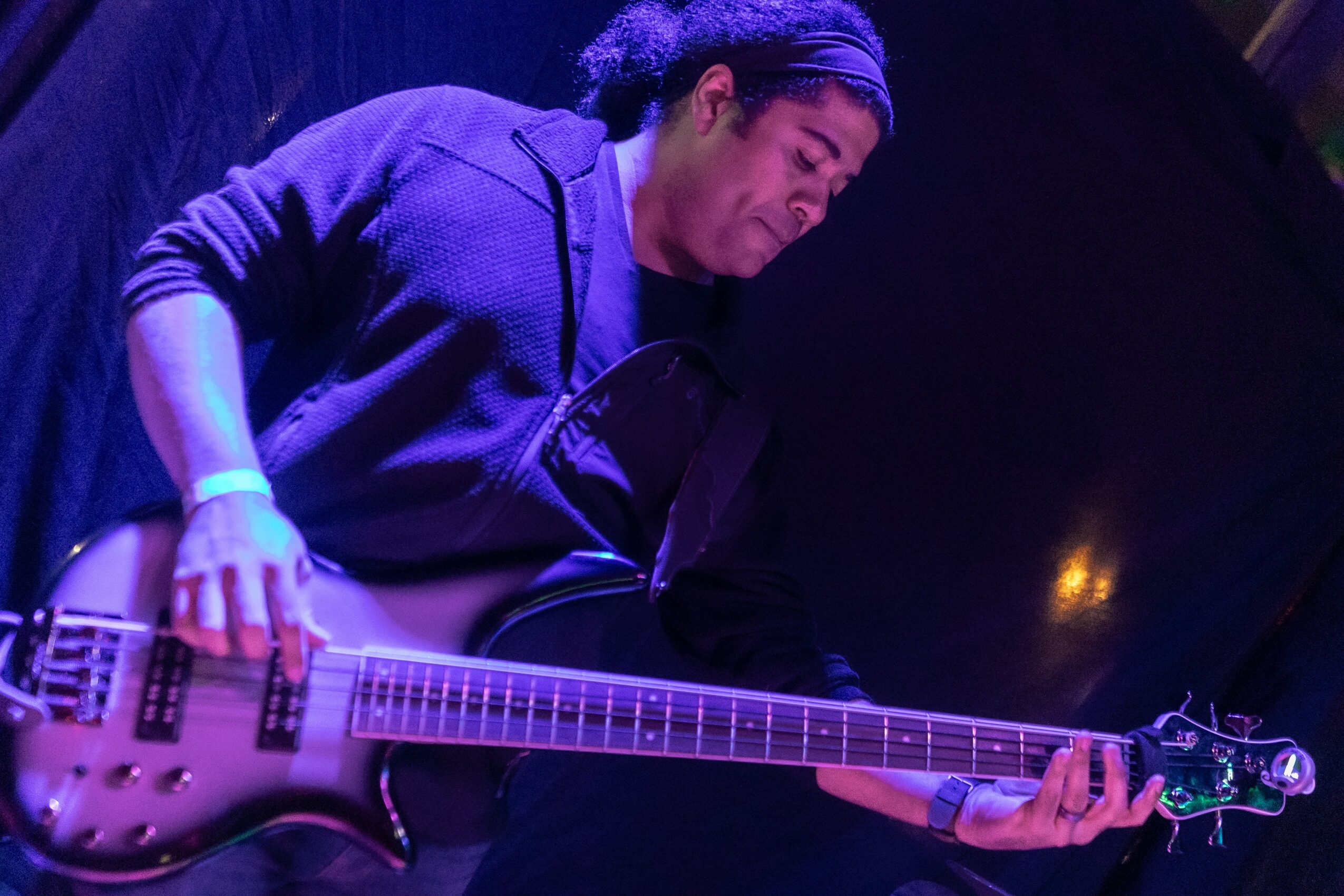 Person playing an electric guitar on stage, wearing a dark hoodie, with blue stage lighting.