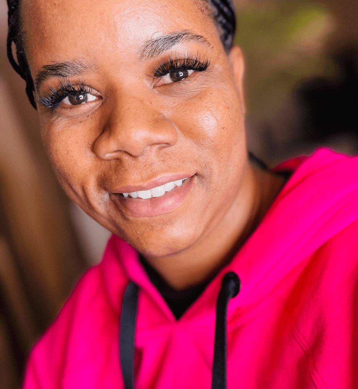 Close-up of smiling woman with dark hair, wearing a bright pink hoodie, outdoors background, natural lighting.
