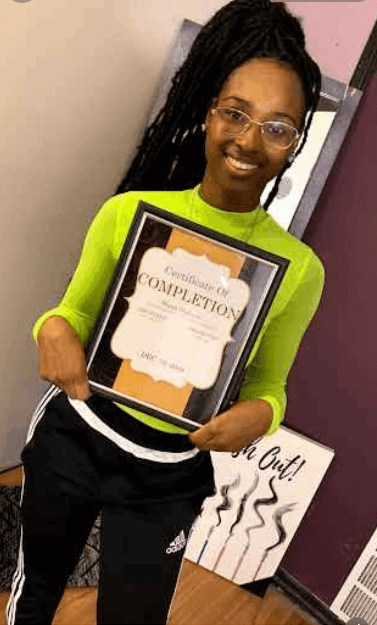 Woman with glasses and braided hair holding a framed certificate, smiling, wearing a bright green top and black pants.