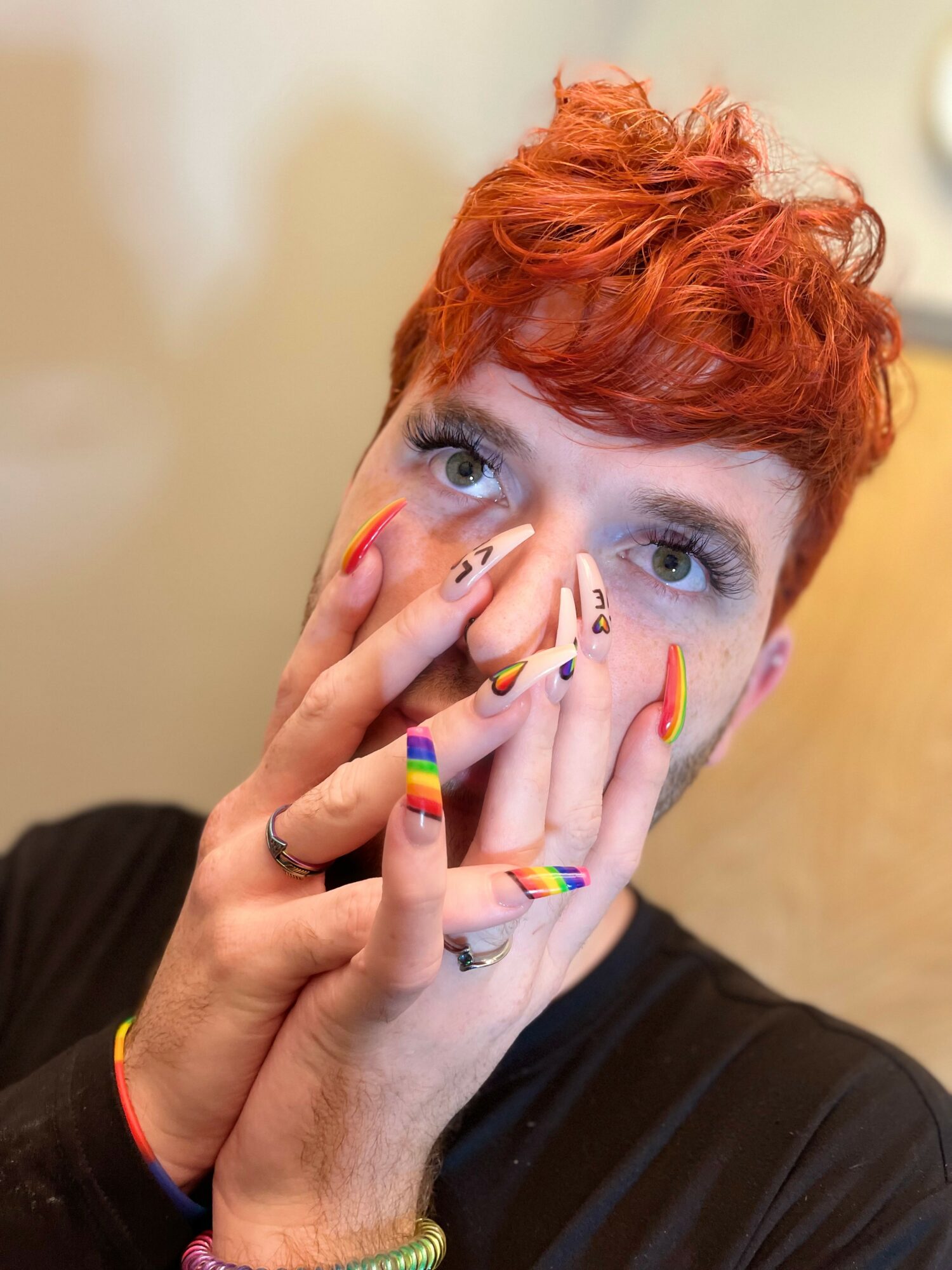 Person with short, curly red hair covering their face with hands, rainbow-colored nails, and a black shirt, against a beige background.