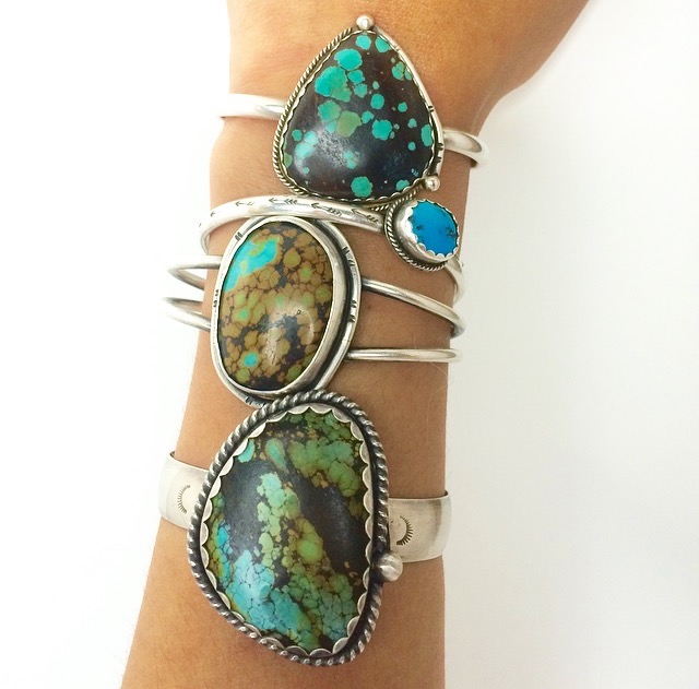 Multiple silver rings with large turquoise and green stones worn on a person's arm.