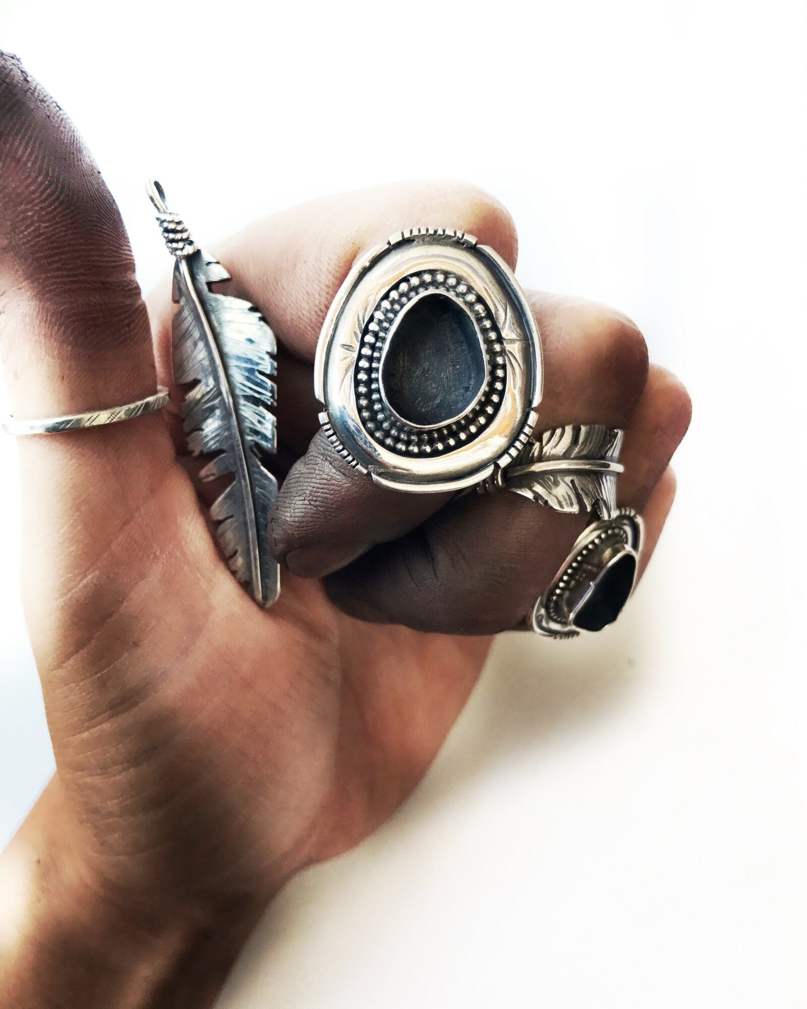 Hand with multiple rings, including a large oval ring, a feather-shaped ring, and a smaller ring, against a white background.