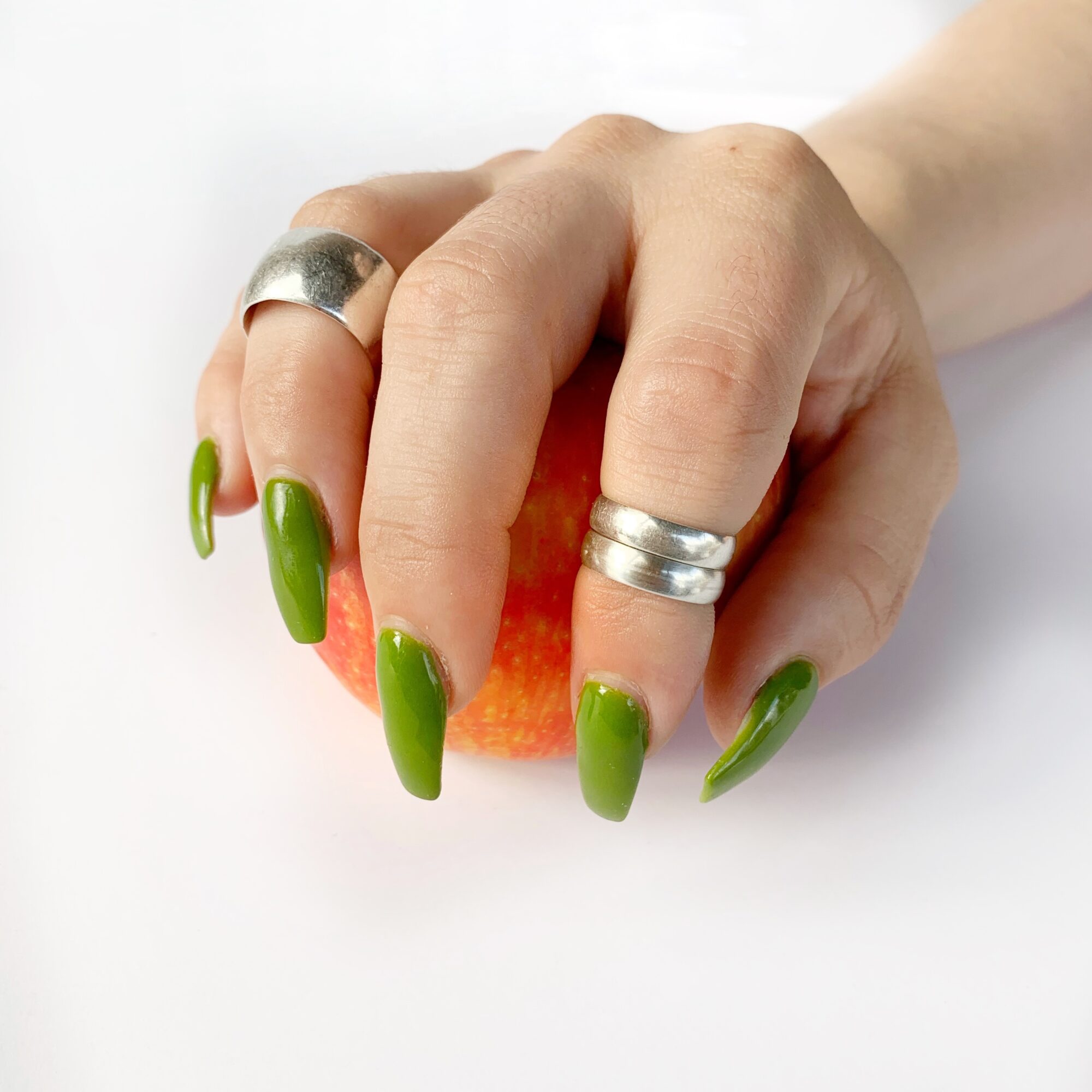 Hand with green nail polish holding an apple, wearing two silver rings, against a white background.