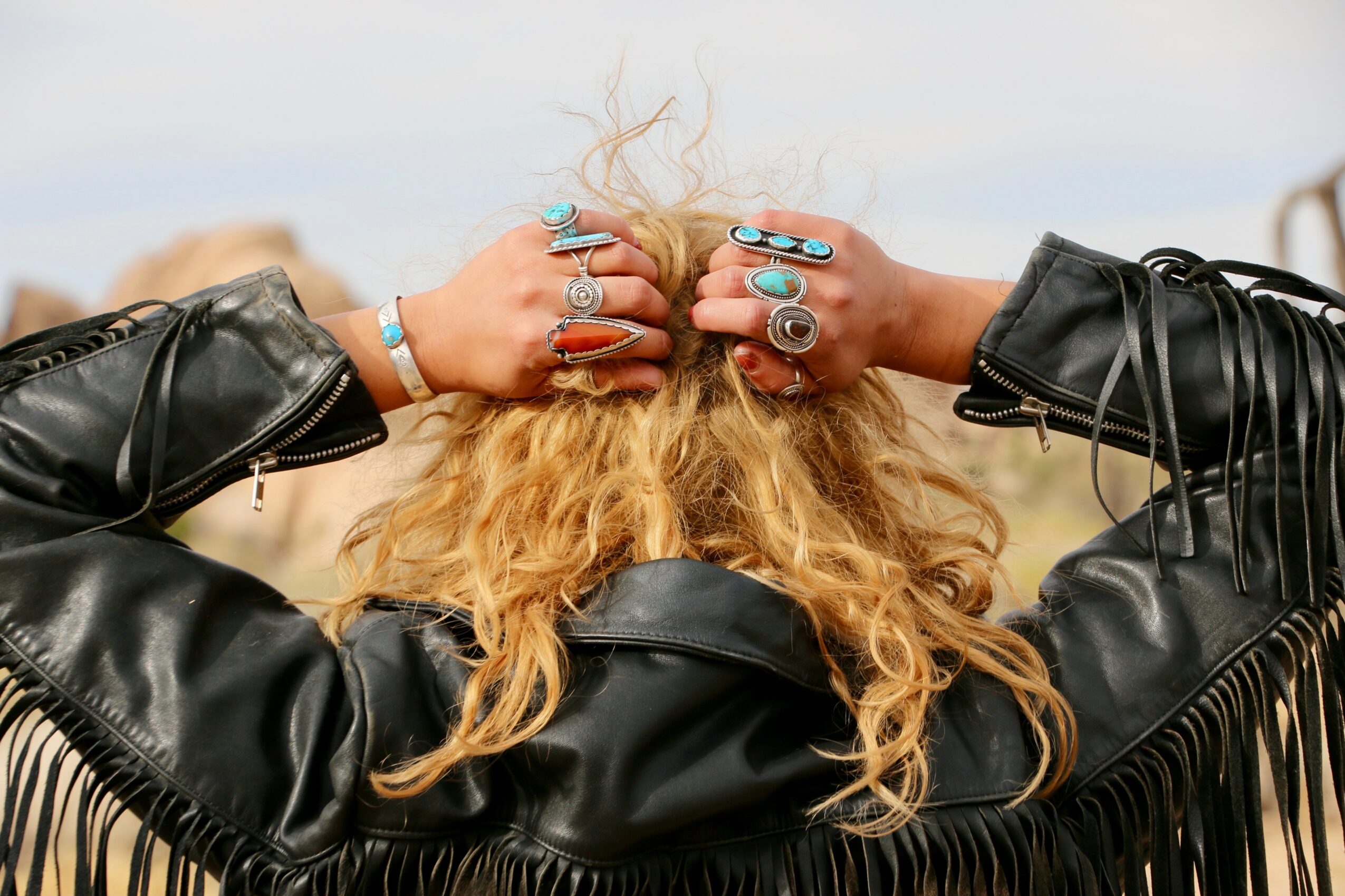 Person with curly blonde hair wearing a black leather jacket, holding their head with both hands, outdoors.