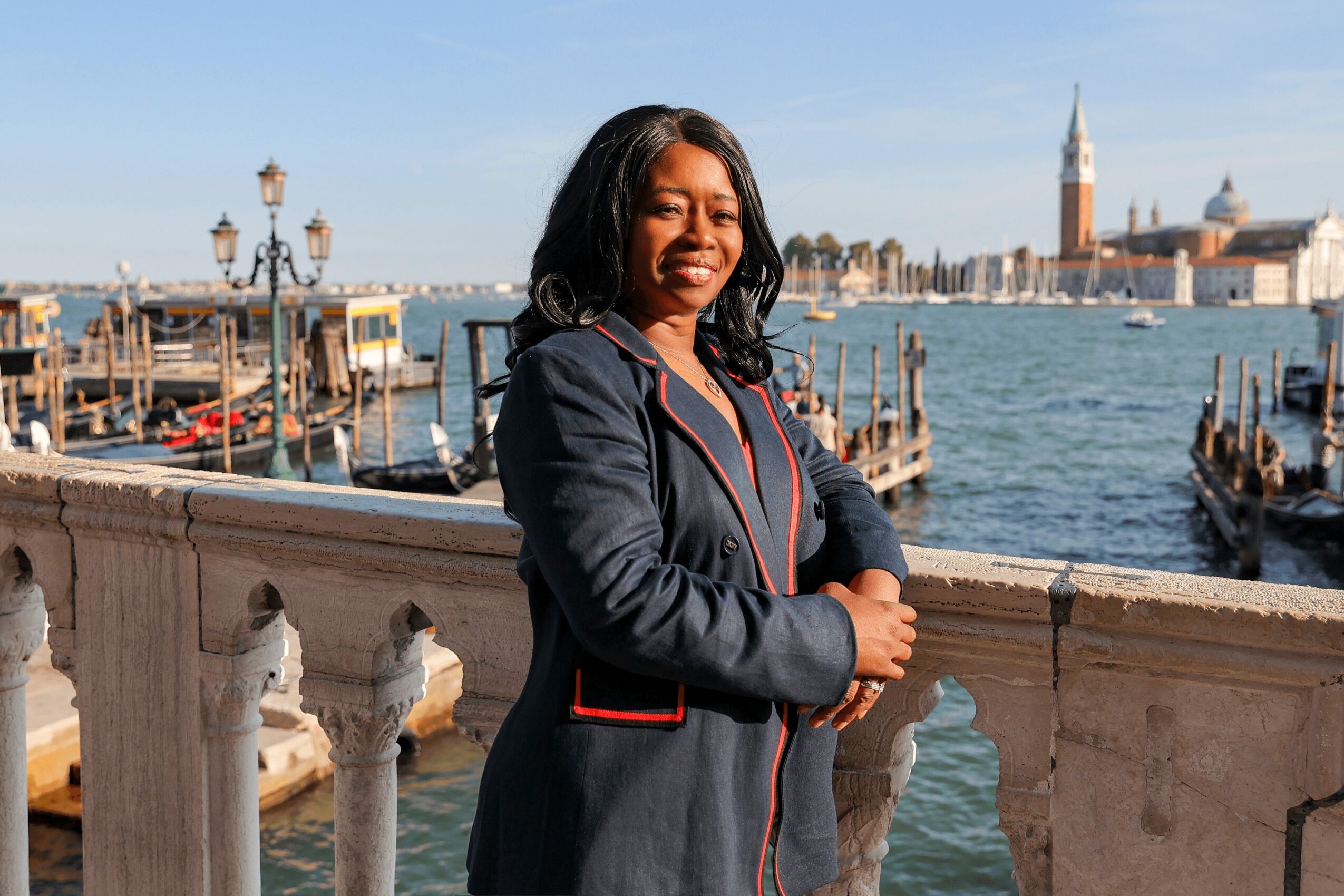 Woman standing by a stone railing near water with boats and historic buildings in the background, smiling.