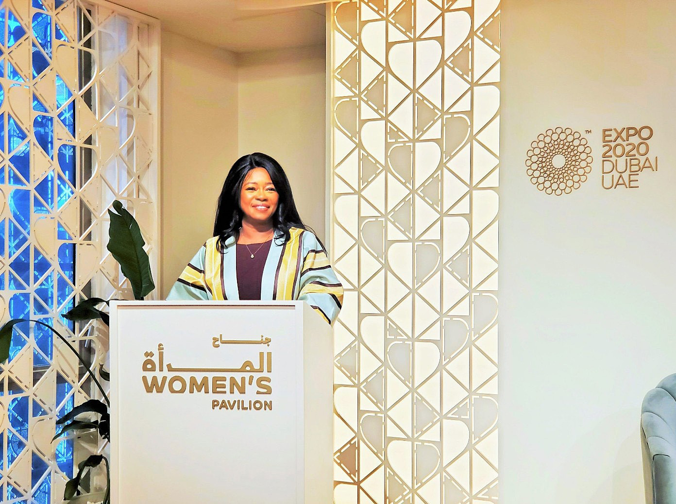 Woman standing behind a podium at Women's Pavilion, Expo 2020 Dubai, smiling, with decorative background and logo.