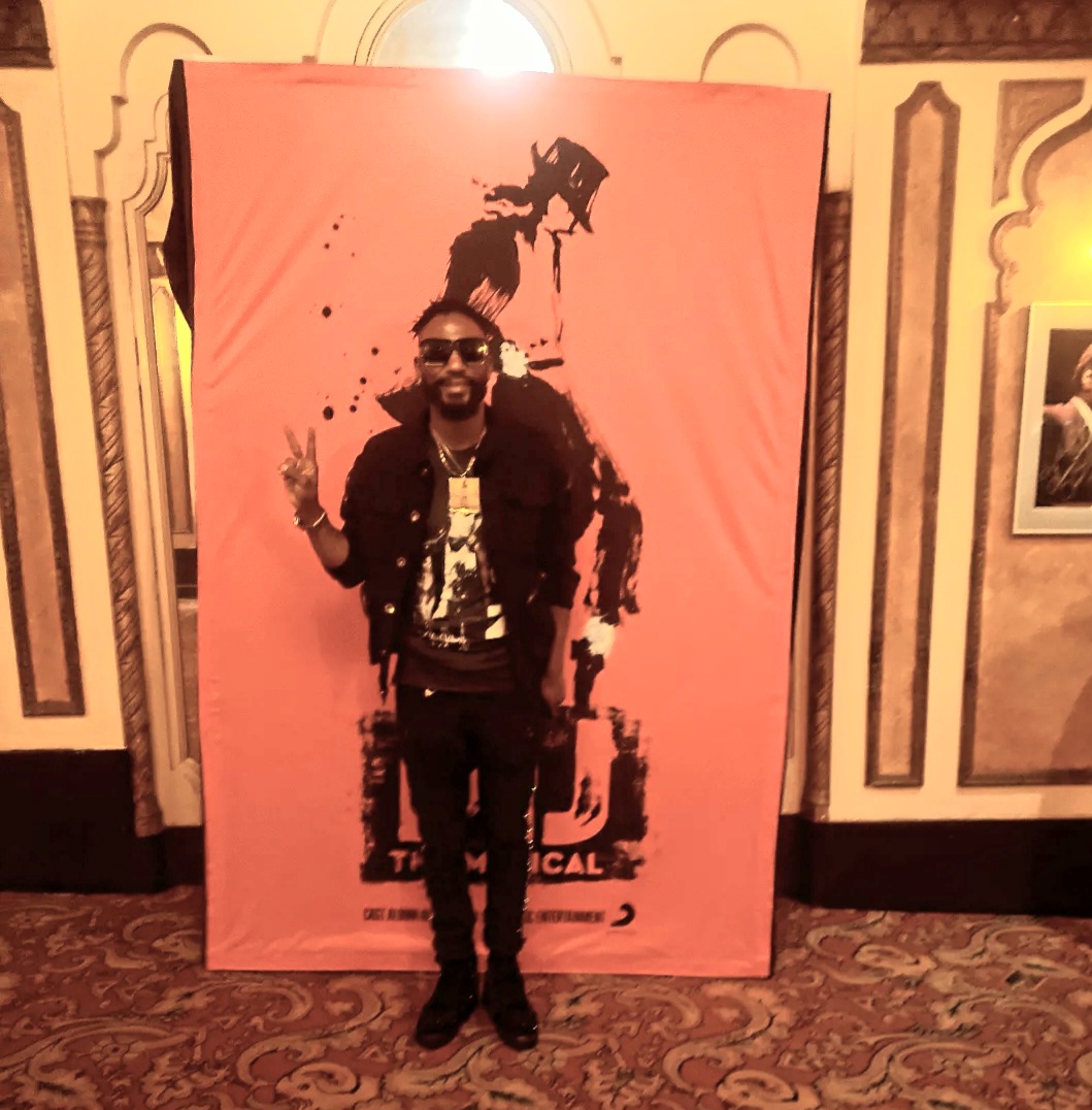 Person standing in front of a pink backdrop with a silhouette of a person, making a peace sign with one hand.