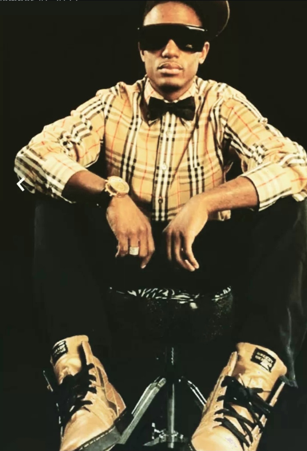 Person wearing sunglasses, hat, plaid shirt, bow tie, sitting with legs apart, hands on knees, against dark background.