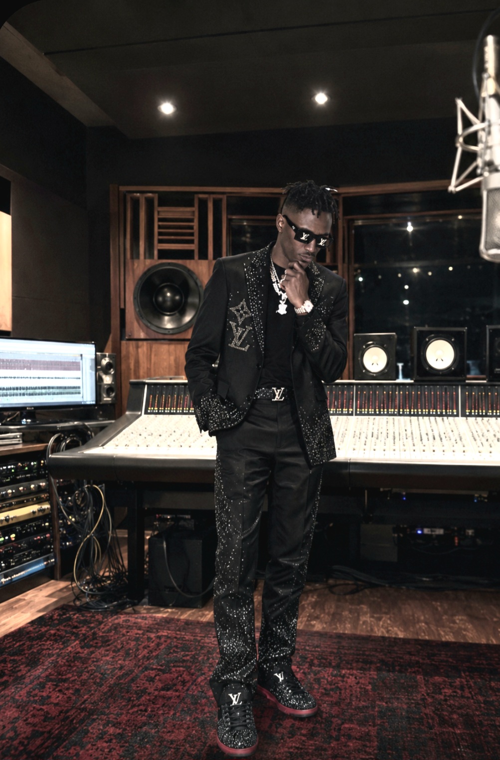 Person in black outfit with jewelry and sunglasses in a recording studio, standing near a large mixing console.