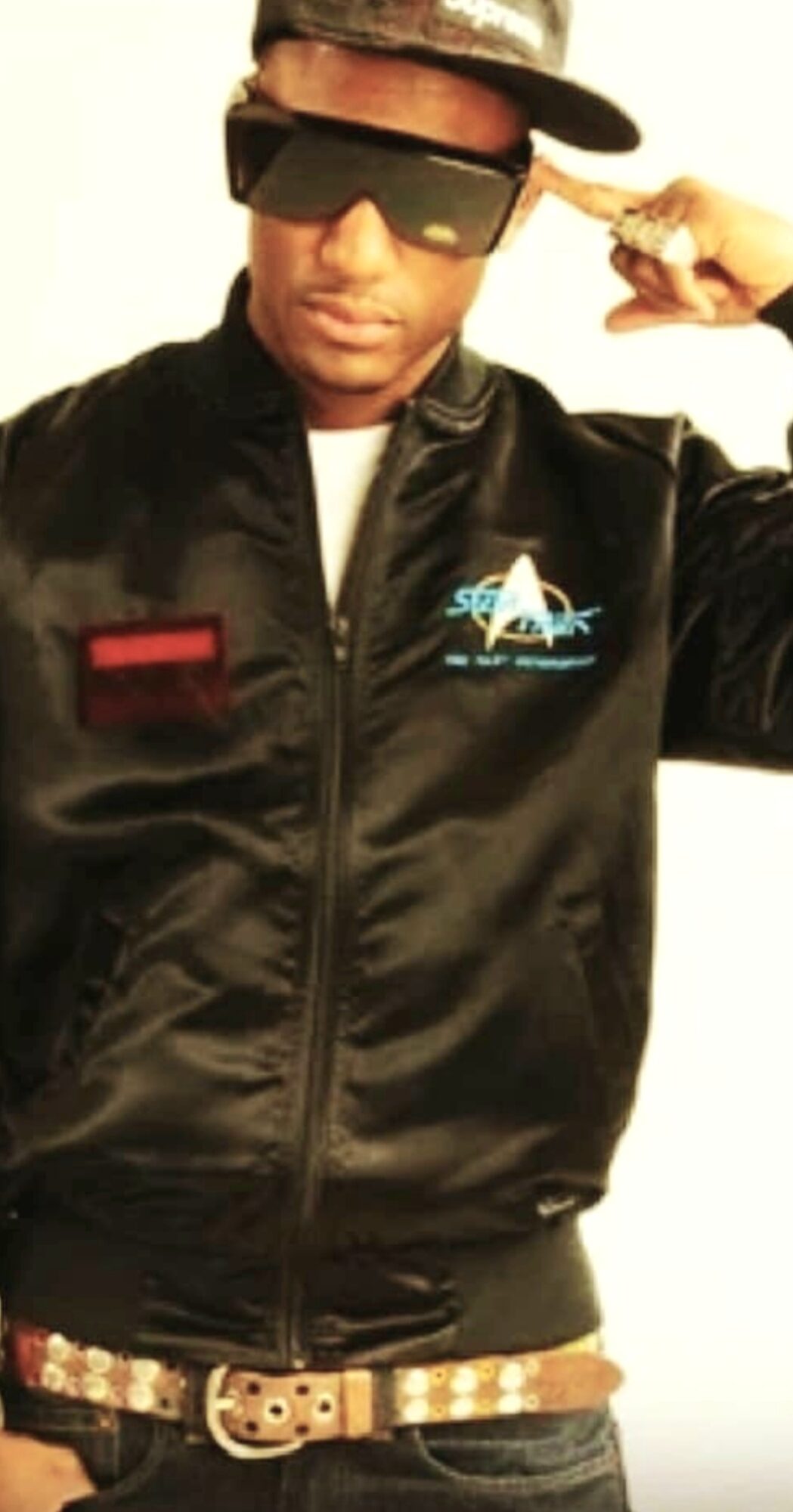 Person wearing sunglasses, a cap, and a black jacket with a logo, pointing to their head.