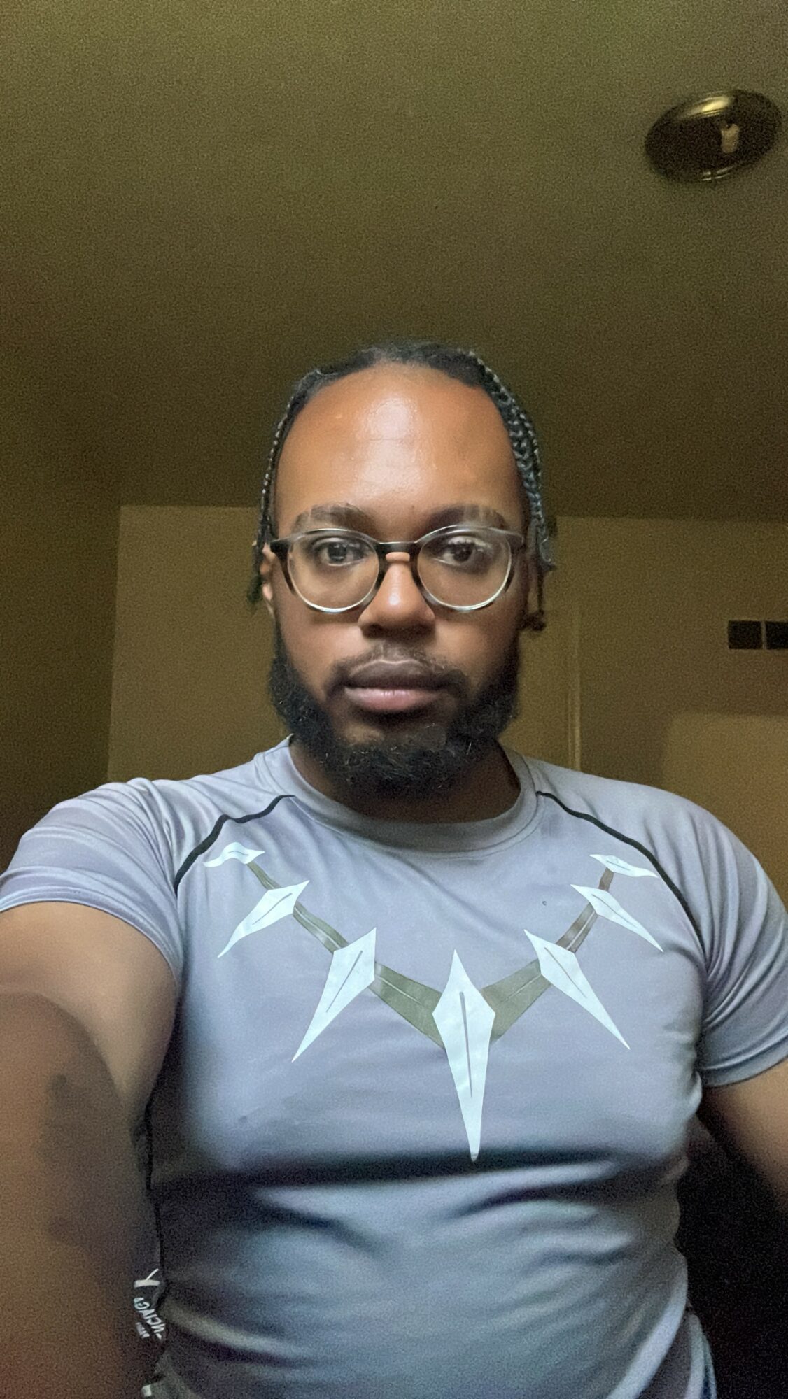 Man with glasses and braided hair taking a selfie indoors, wearing a gray shirt with geometric design, neutral expression.
