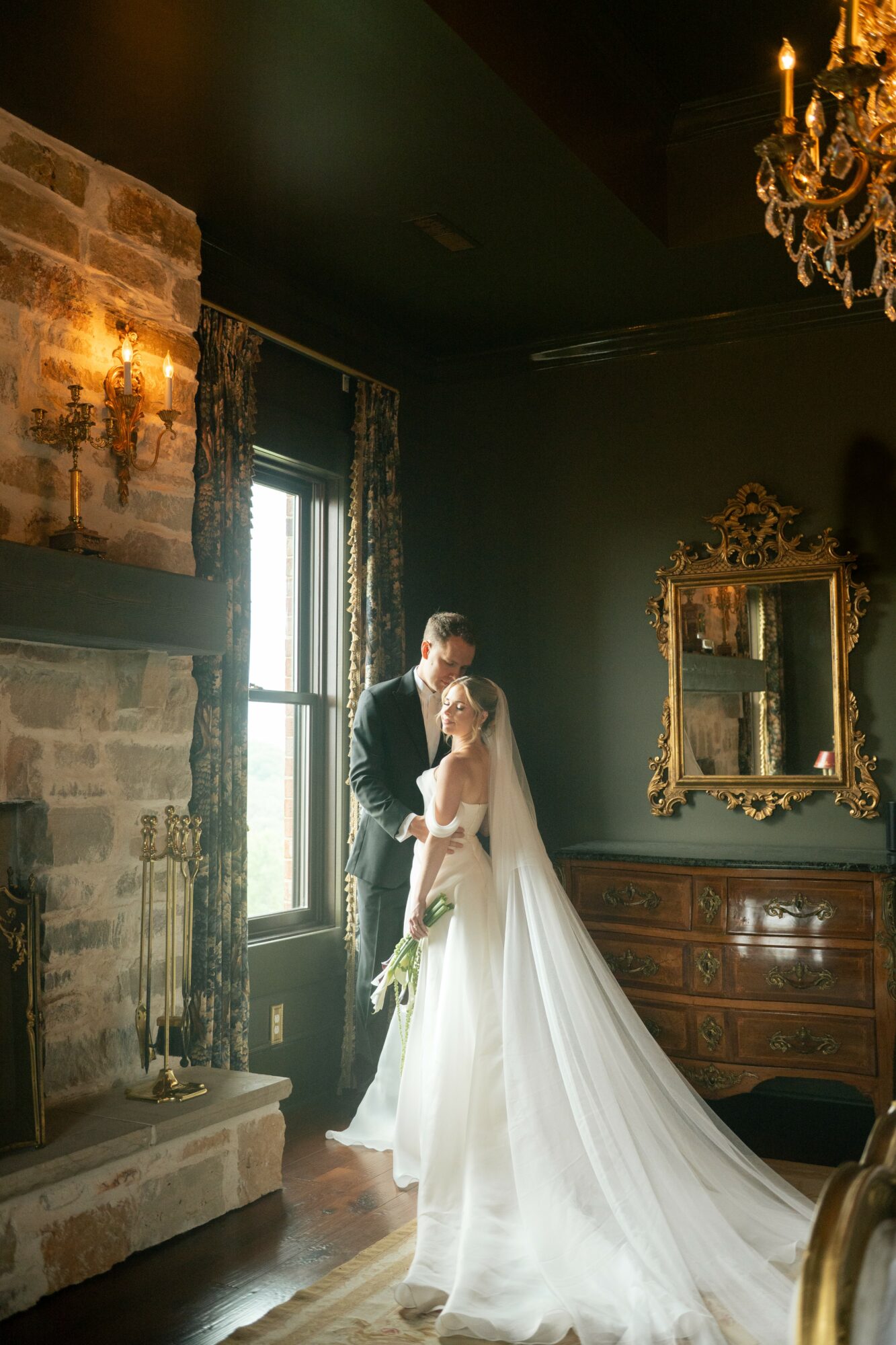 A bride and groom stand together indoors near a window, with a chandelier and ornate mirror in the background.