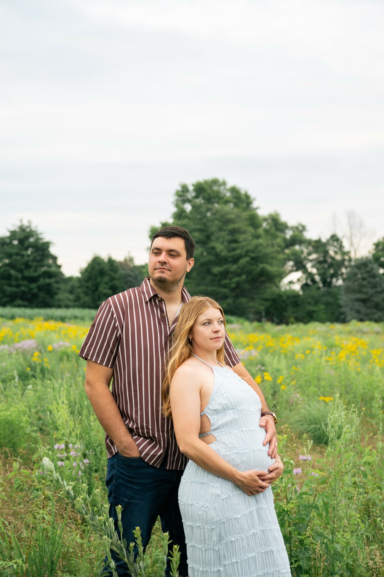 A man and pregnant woman standing in a field with trees and yellow flowers, the woman holding her belly.