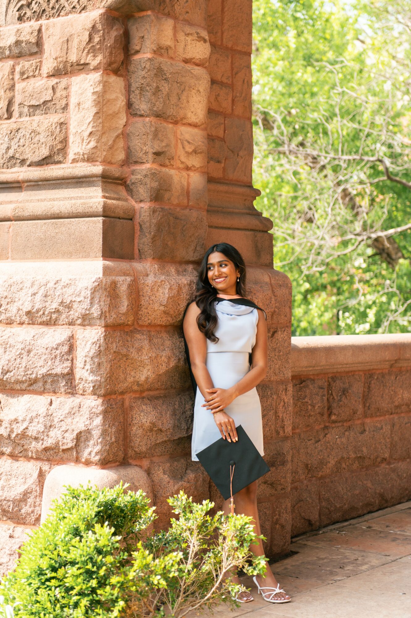Woman in a light blue dress standing against a stone wall outdoors with green trees in background.