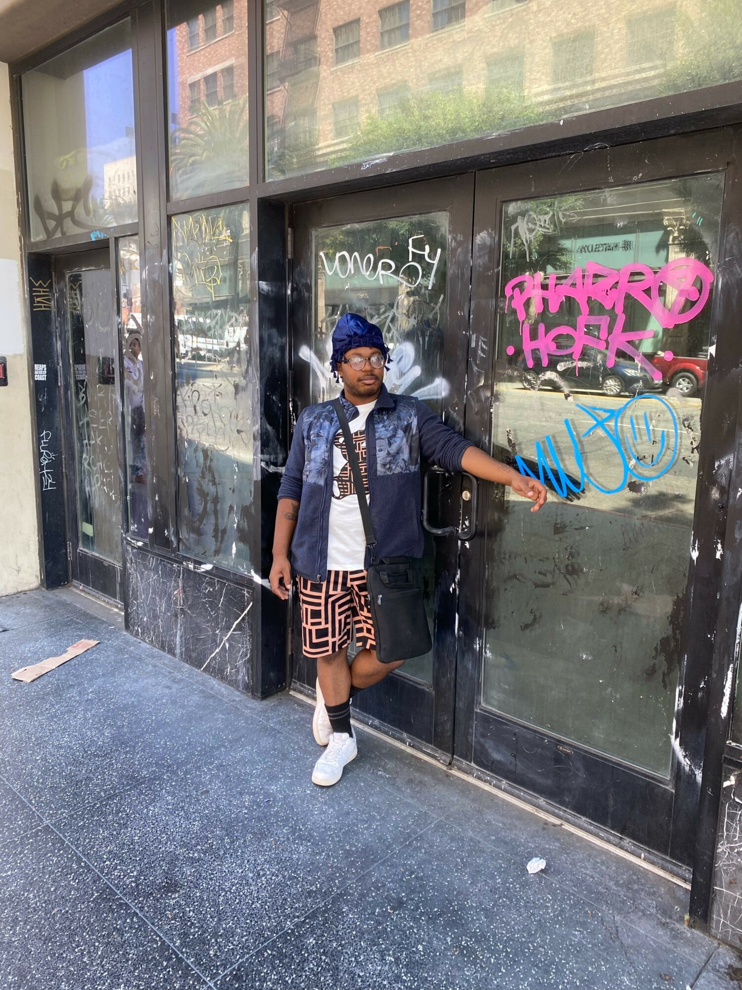 Person standing outside a glass door with colorful graffiti, wearing a denim jacket, shorts, and white shoes, holding a phone.