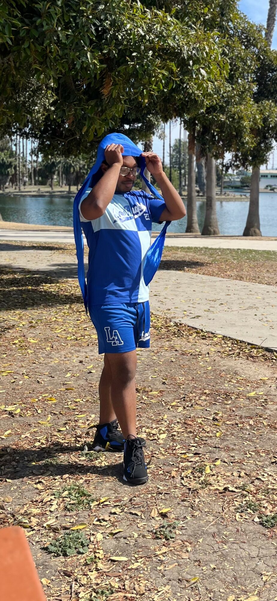 Child in blue sportswear holding a blue ball above head outdoors near water and trees.