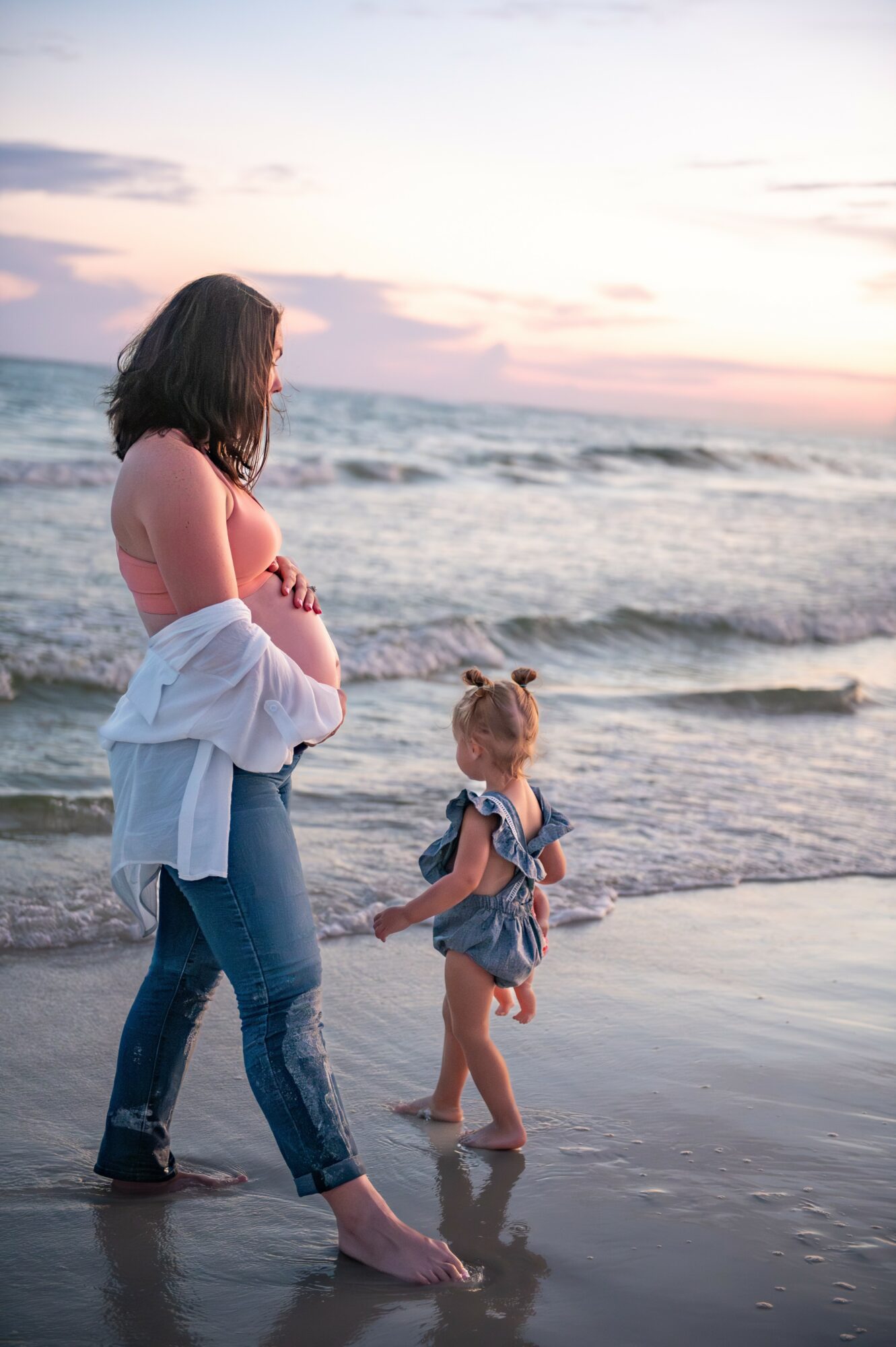 Woman and child standing in shallow ocean water at sunset, facing each other, woman with arms crossed, child holding hand, waves in background