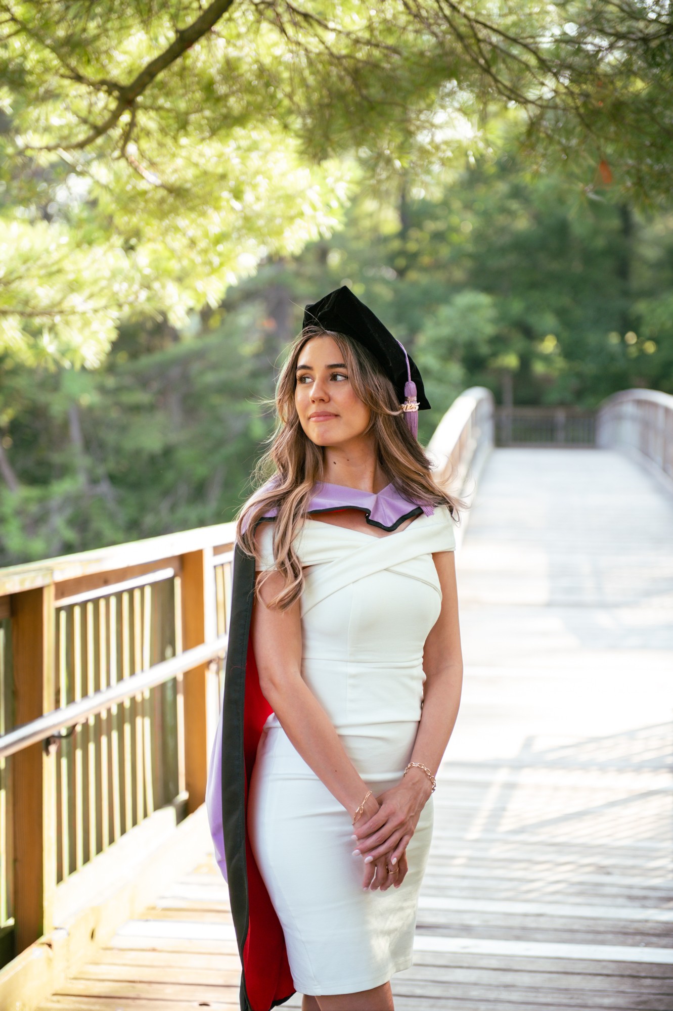 Woman in a white dress with a purple and black graduation gown on a bridge outdoors, trees in background.