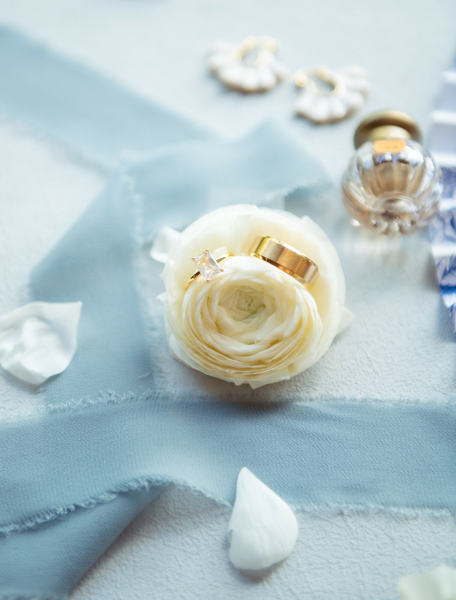 Flat lay of perfume bottles, a flower, and petals on a light blue fabric surface.