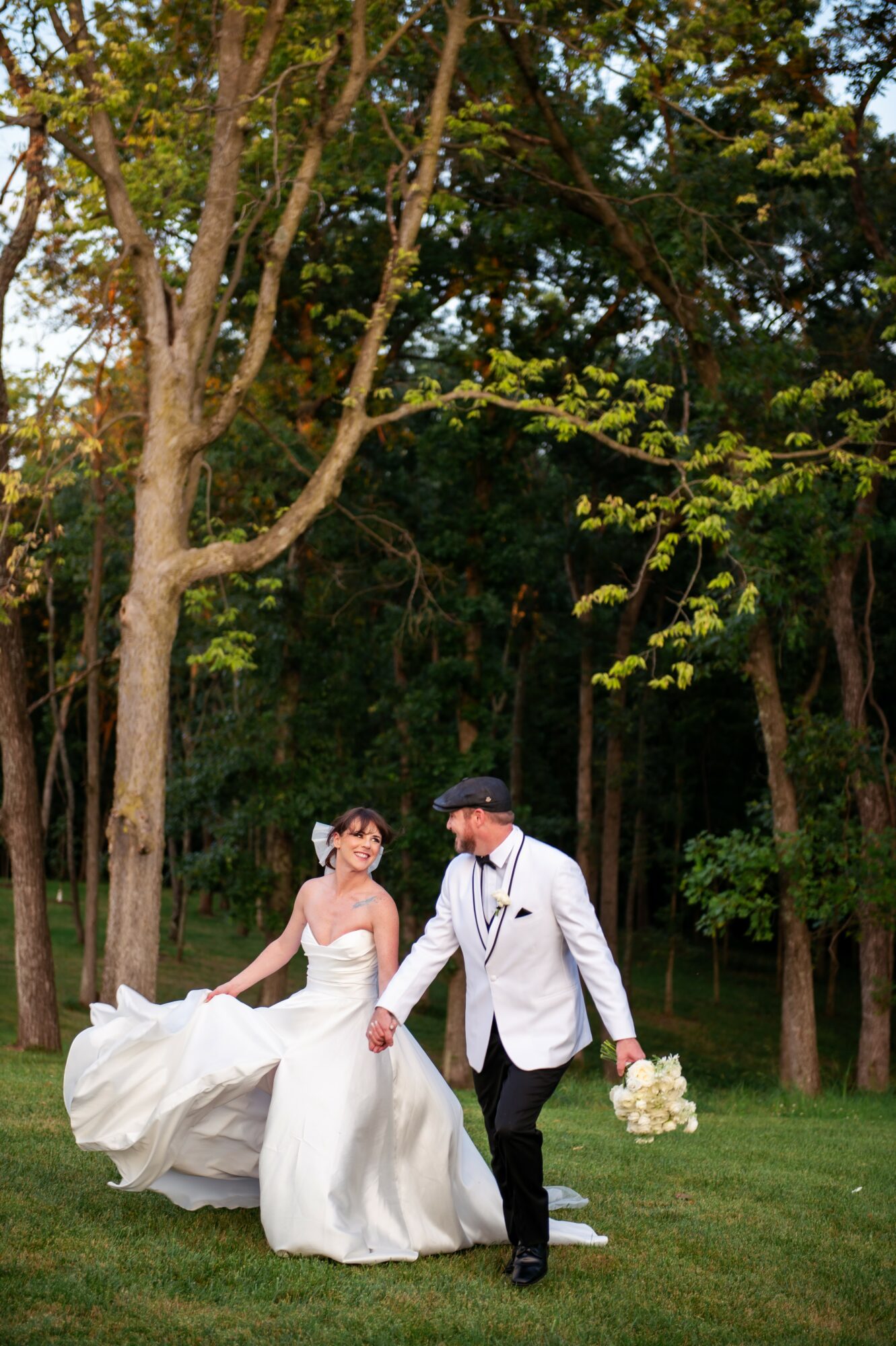 A bride and groom walk hand in hand outdoors, surrounded by trees and greenery, holding a bouquet of flowers.
