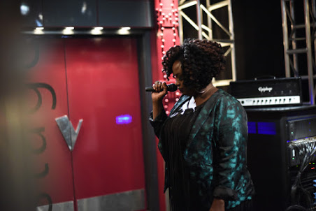 Person with curly hair holding a microphone, standing near a red door and audio equipment in a stage setting.