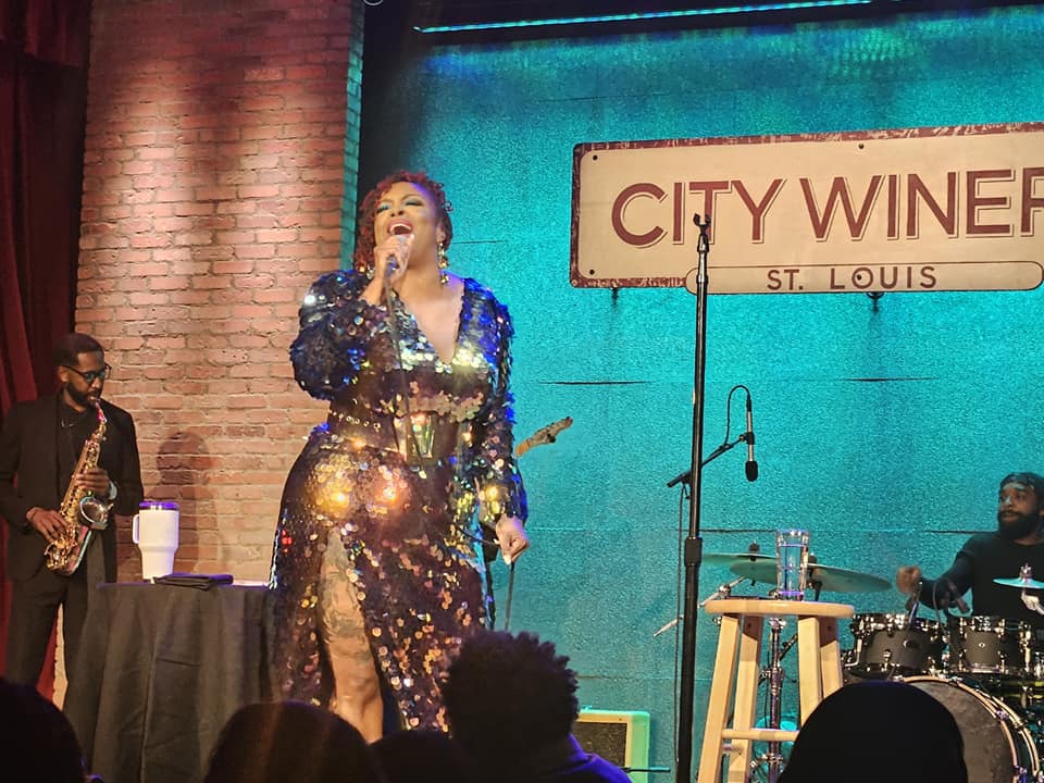 Singer performing on stage with a saxophonist and drummer, brick wall background, city winery sign, and audience visible.