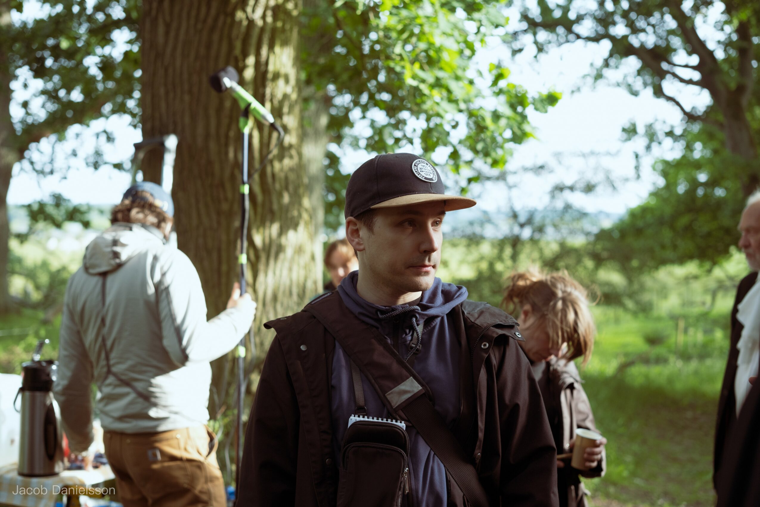 Young man wearing a black cap and jacket outdoors with trees and greenery in background.
