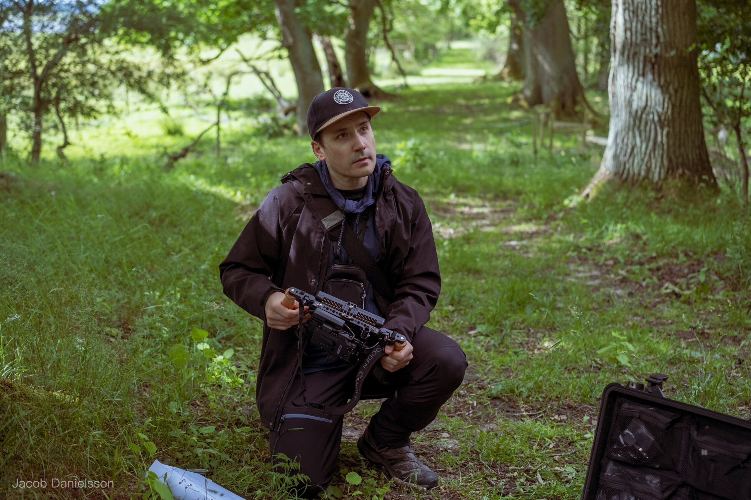 Person crouching in a forest holding a black object, with a case open on the ground nearby, surrounded by trees and greenery.