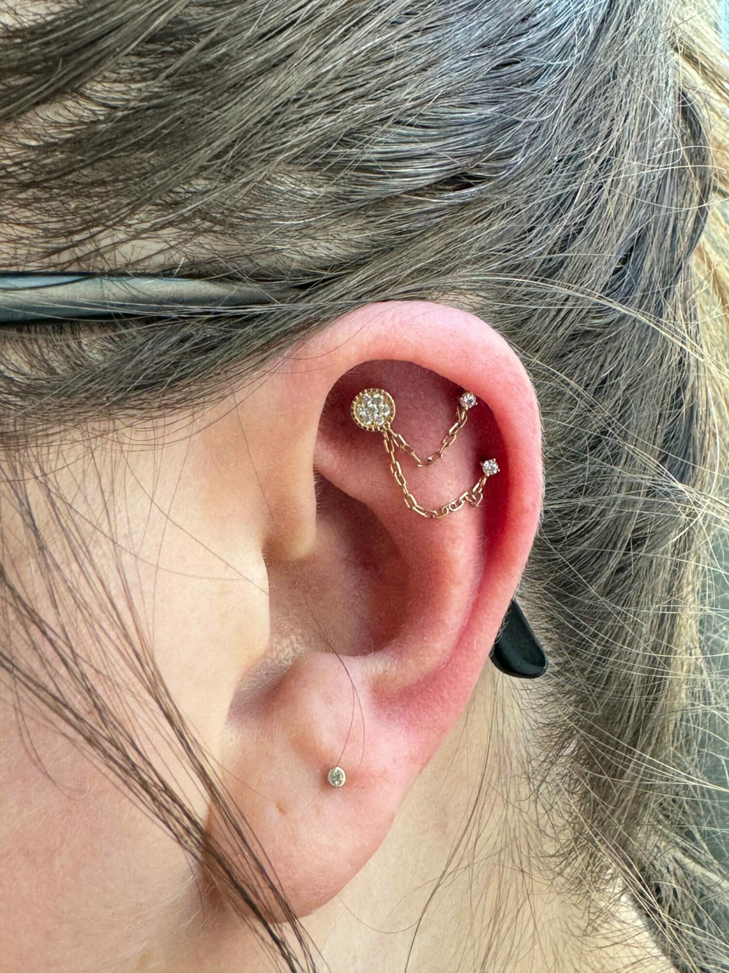 Close-up of a person's ear with a gold chain and pendant earring, and a small stud earring, with hair partially covering the ear.
