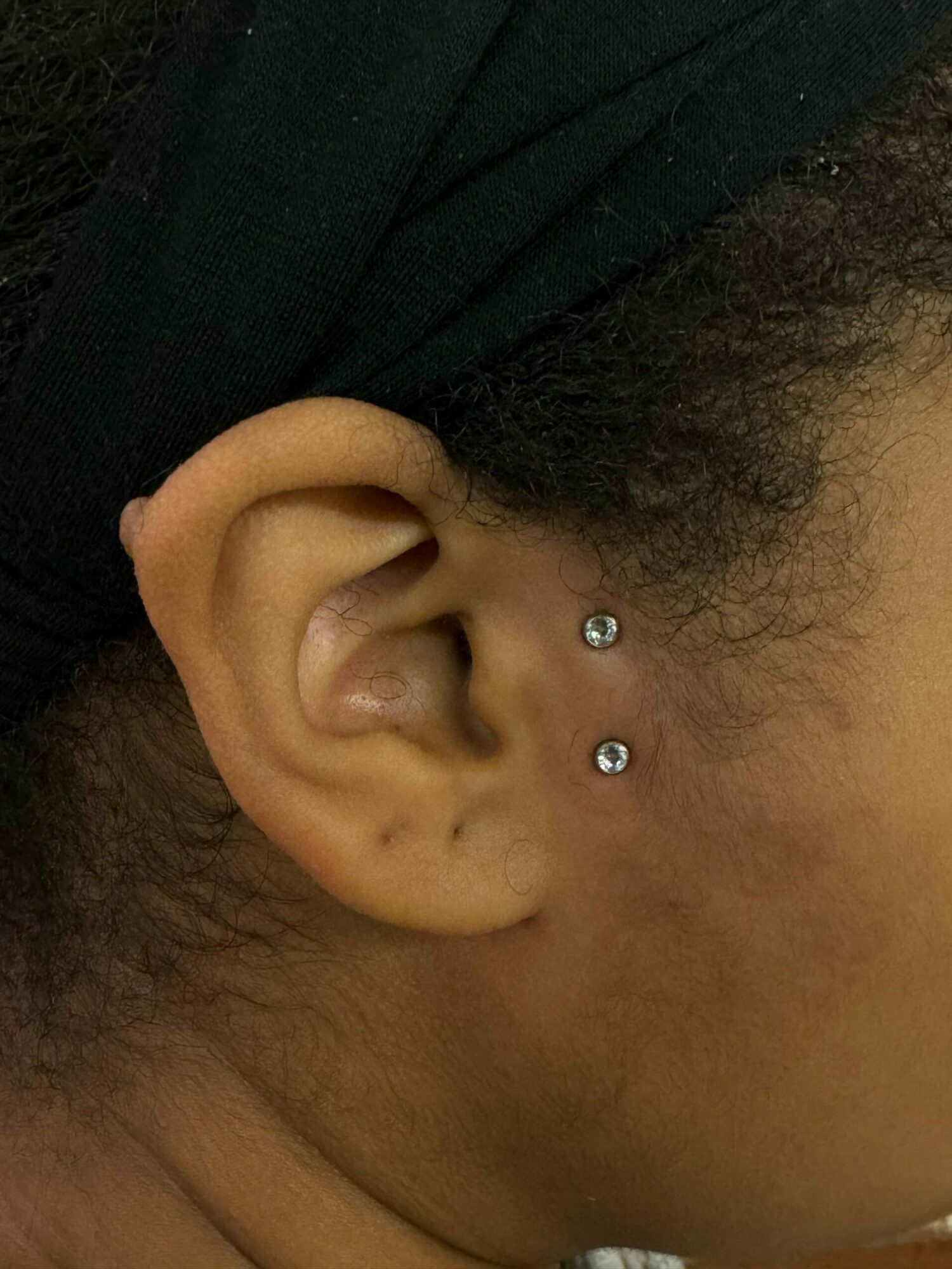 Close-up of a person's ear with two small piercings, surrounded by dark hair and skin.