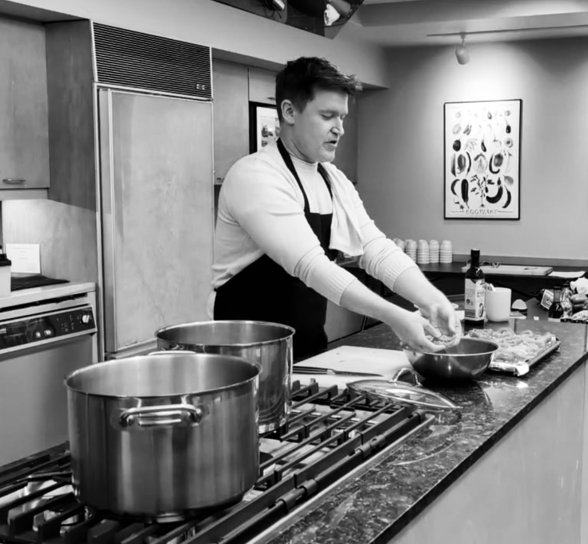 Young man cooking in a kitchen, wearing an apron, with pots on the stove and utensils on the counter.