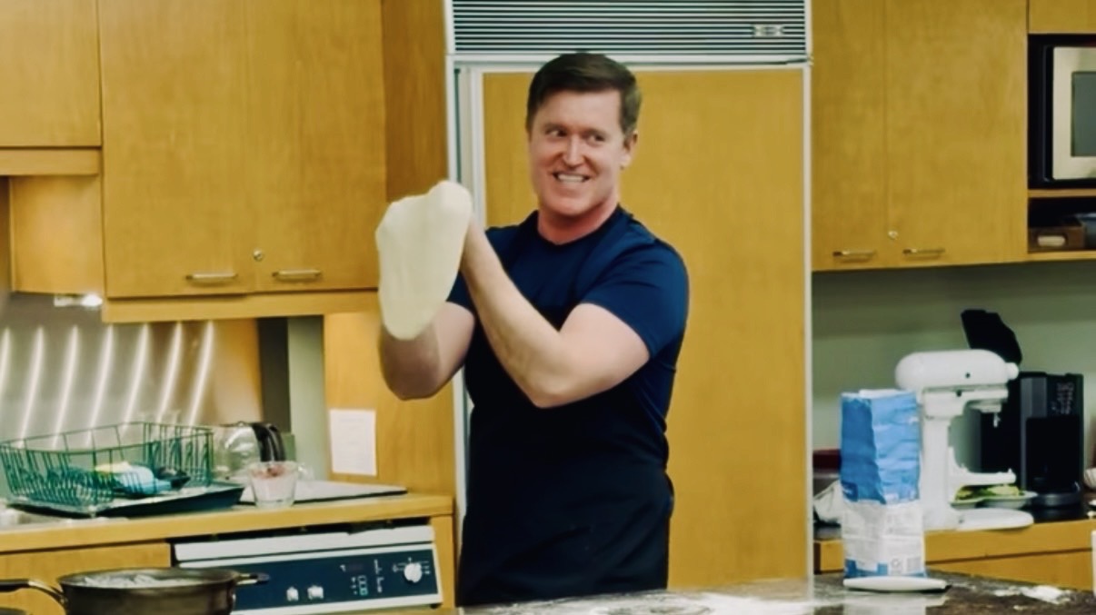 Man in a kitchen holding a white cloth, smiling, with wooden cabinets and kitchen appliances in the background.