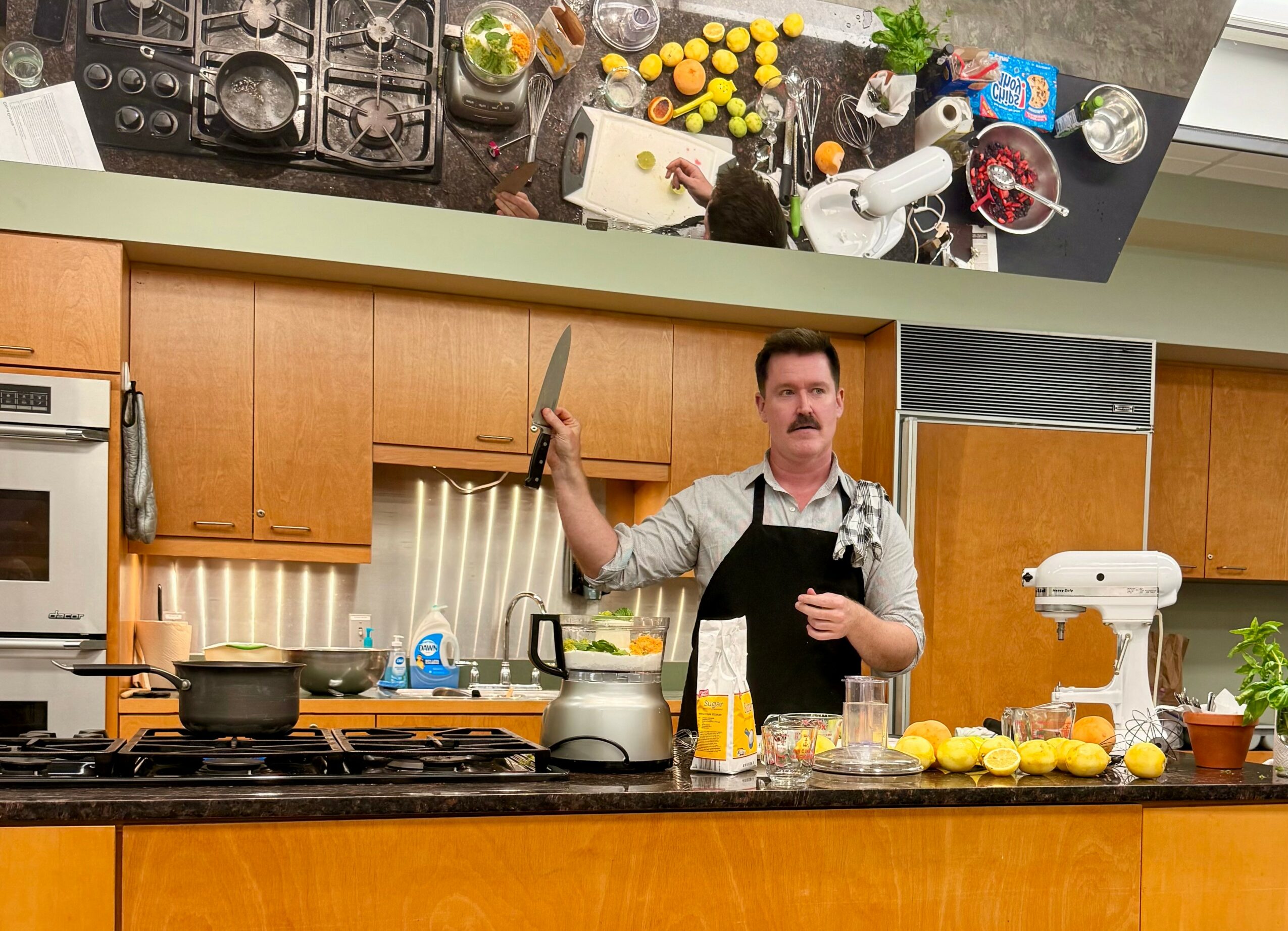 Man in apron holding a knife in a kitchen with wooden cabinets, stove, and various cooking utensils and ingredients on the counter.